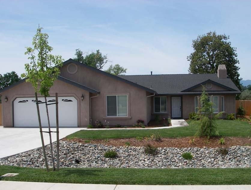 Single-story suburban house with a two-car garage, a small front yard with trees and landscaping, and a concrete driveway under a clear blue sky.