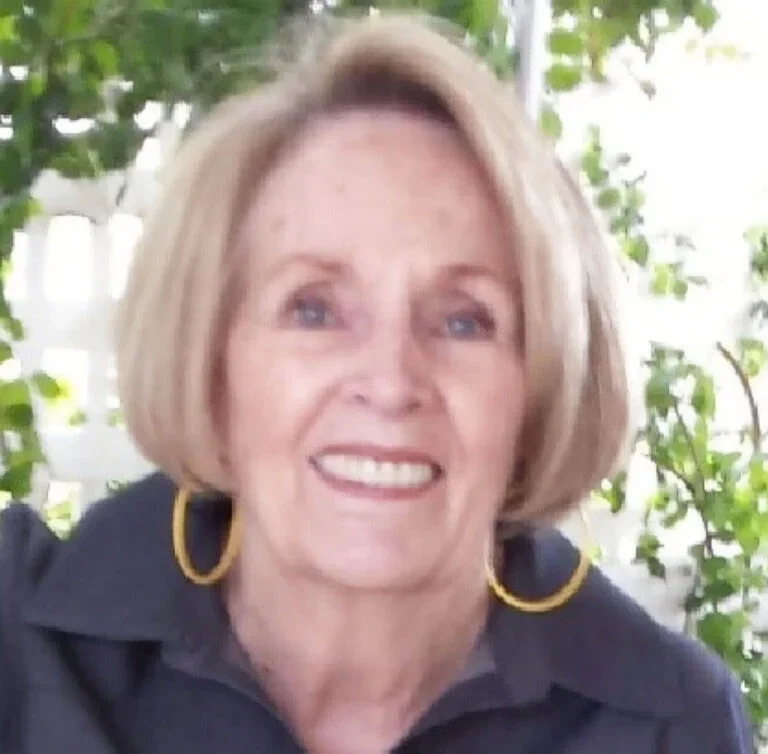 A woman with shoulder-length blonde hair, smiling, wearing yellow hoop earrings and a dark top, with greenery in the background.