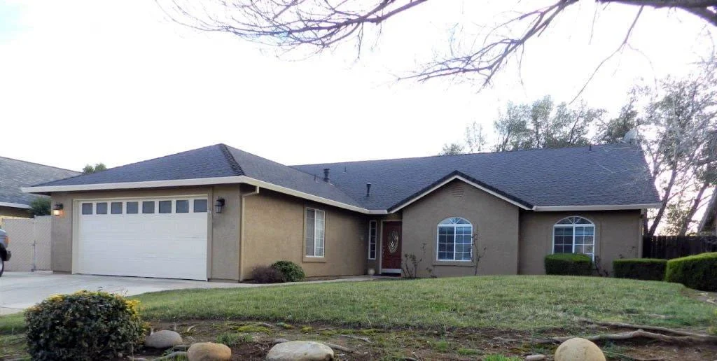 Single-story house with a beige exterior, white garage door, black roof, front yard with grass, bushes, and rocks, and a tree in the background.