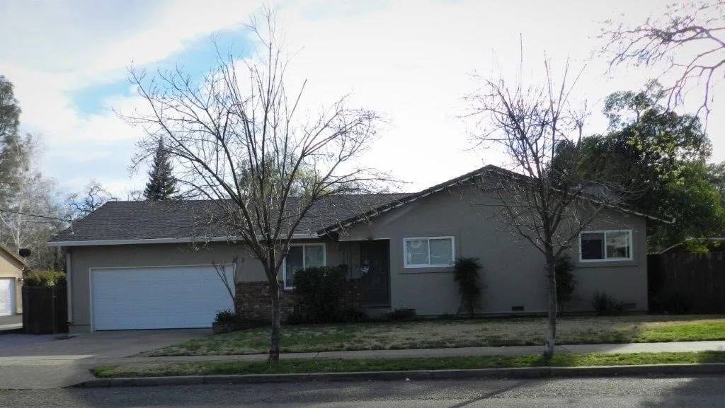 A single-story suburban house with a gray exterior, two leafless trees in the front yard, and a garage with a white door. The sky is partly cloudy.