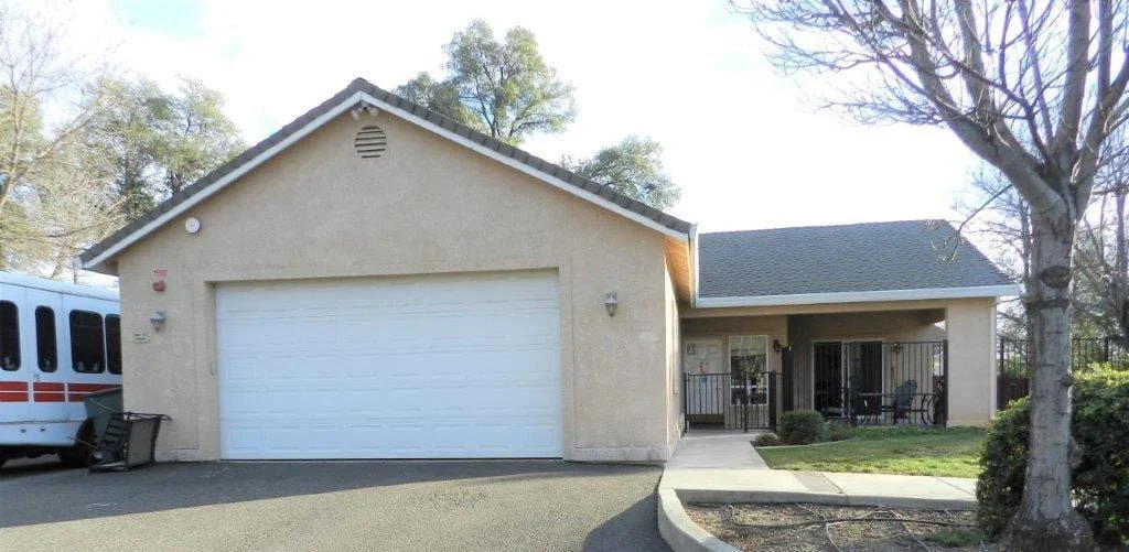 A beige house with a white garage door, a small porch with seating, and a tree in the front yard.