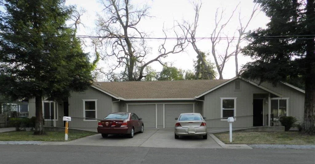 A single-story house with two cars parked in the driveway, surrounded by trees and a grassy lawn, with mailboxes in front.