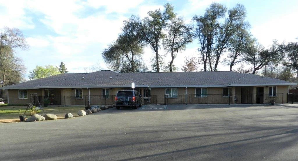 Single-story apartment building with a black vehicle parked in front, surrounded by a paved parking lot and trees in the background.