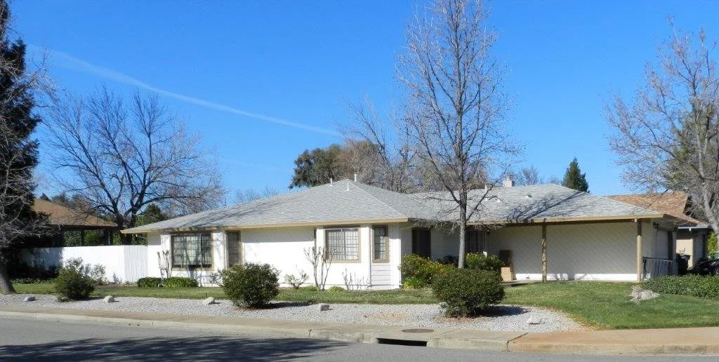Single-story white house with a gray roof, surrounded by trees and shrubs, with a front yard and a sidewalk in front, under a clear blue sky.