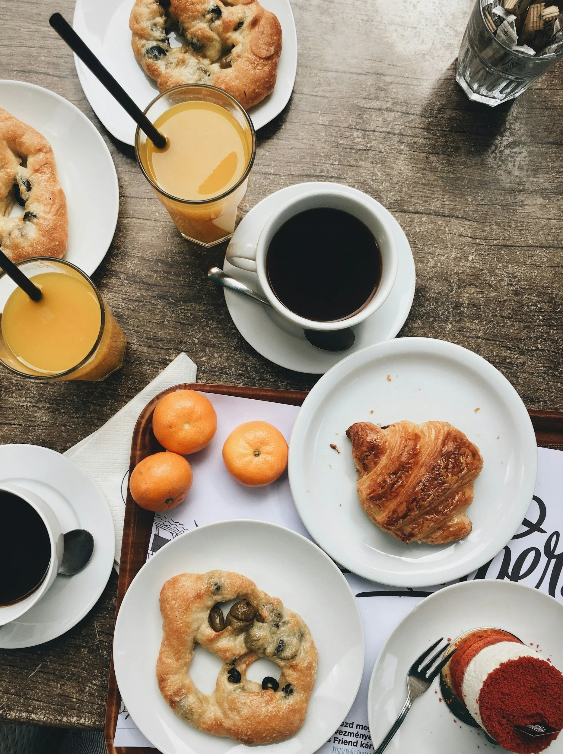 A table set with various breakfast items including a croissant, a muffin with blueberries, a croissant with chocolate, a slice of layered cake with red and white layers, a cup of black coffee, glasses of orange juice with straws, a bowl of black coffee, a plate with oranges, and a glass filled with sugar packets.