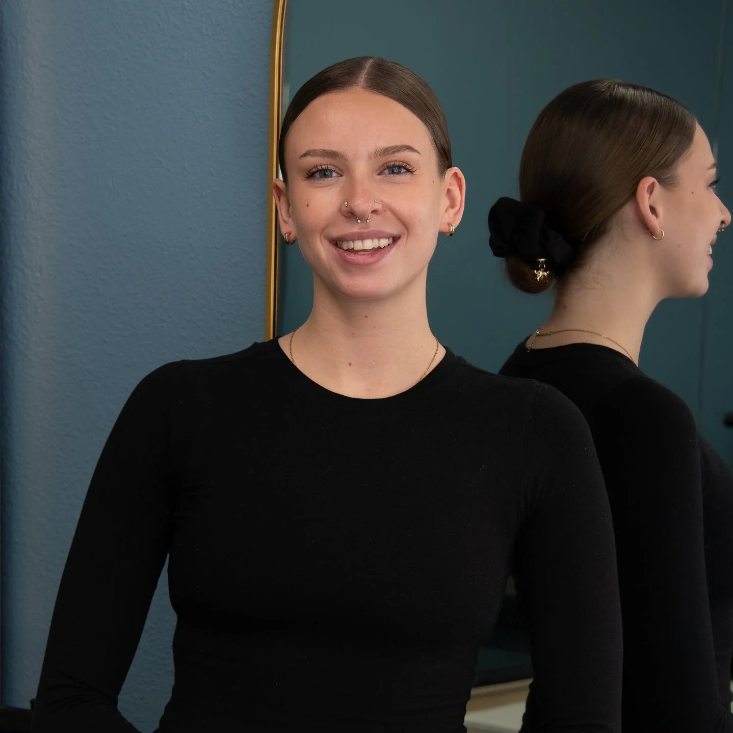 Portrait of a smiling young woman with dark brown hair tied in a bun, wearing a black top, standing next to a mirror reflecting her profile.