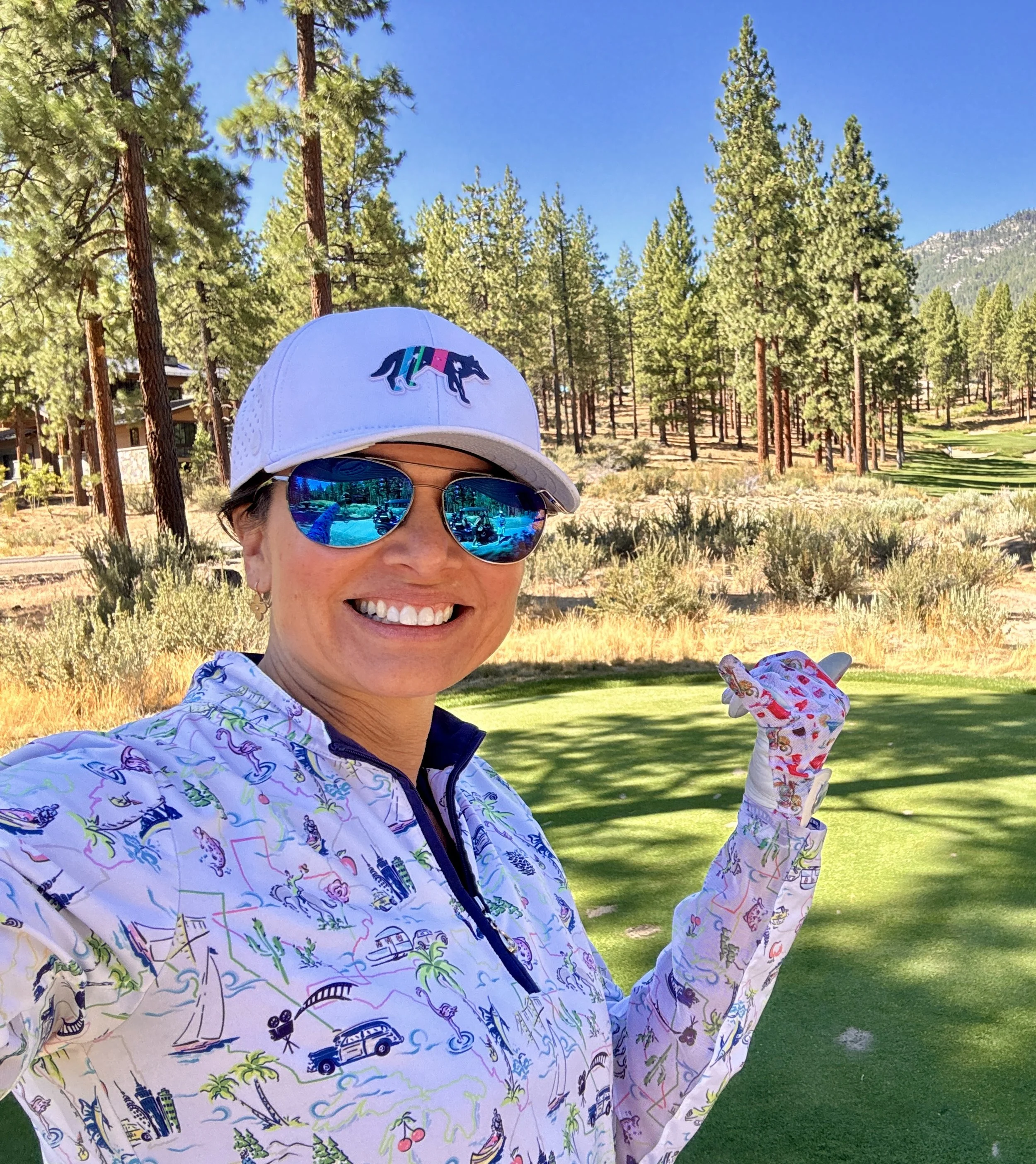 A woman smiling on a golf course, wearing a white cap with a colorful logo, mirrored sunglasses, a white jacket with colorful outdoor-themed prints, and gloves. She is holding a golf tee with a golf ball, surrounded by trees and a green field under a clear blue sky.