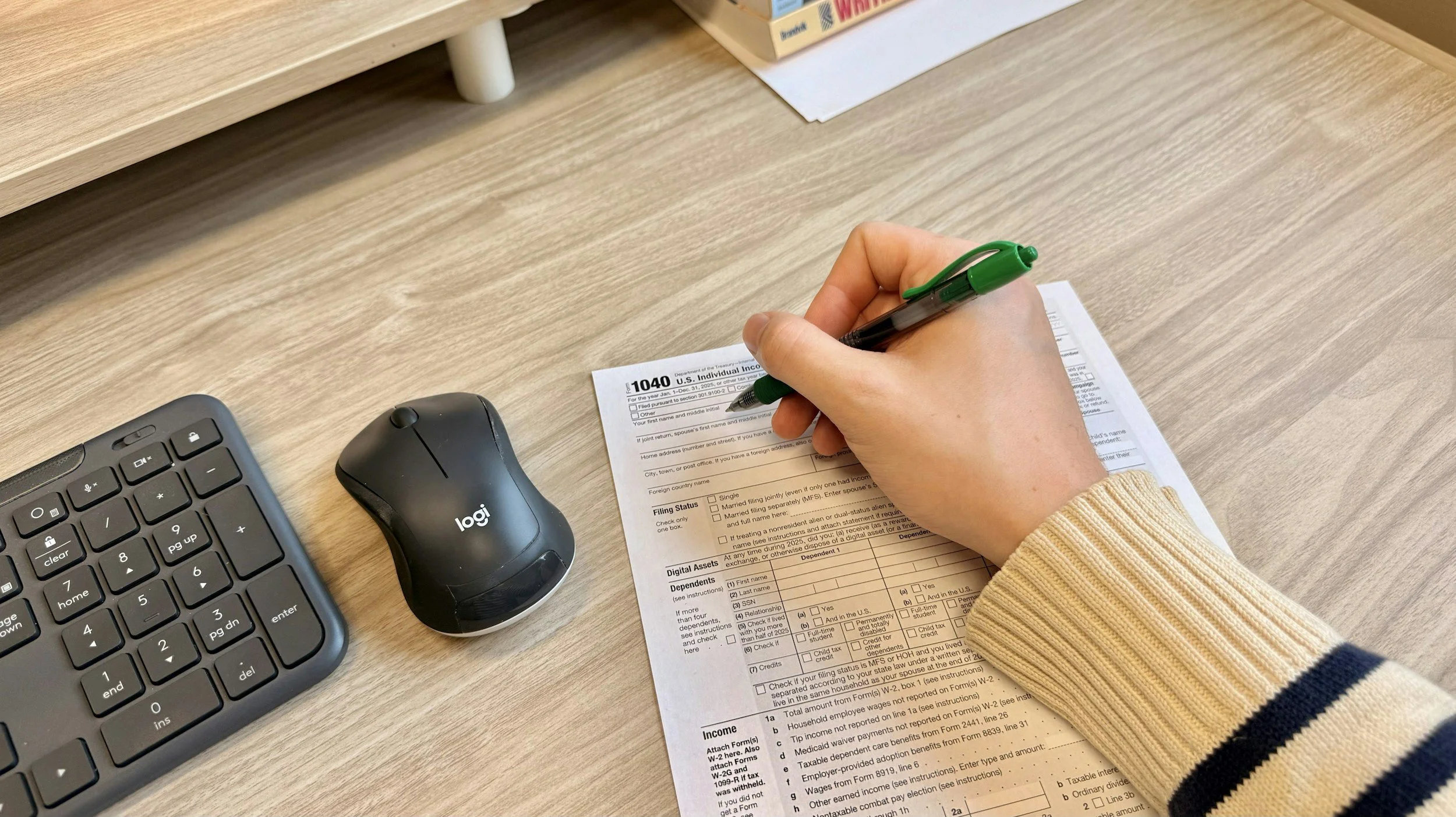 A person filling out a tax form at a desk. The form is a U.S. tax document, the person's hand is holding a green pen, and there is a keyboard and a Logitech mouse nearby.
