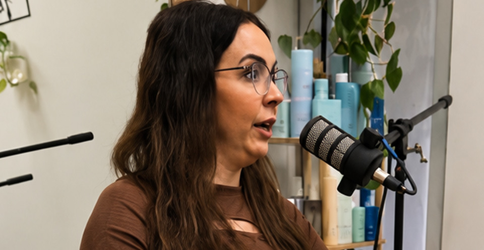 Woman wearing glasses speaking into a studio microphone, with hair products and a houseplant in the background