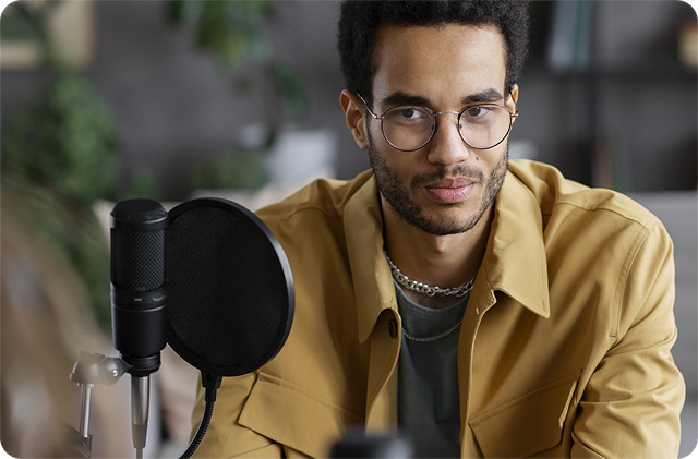 Man wearing glasses and a mustard jacket speaking into a studio microphone with a pop filter