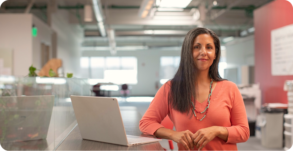Professional woman in a coral sweater standing beside an open laptop in a modern office