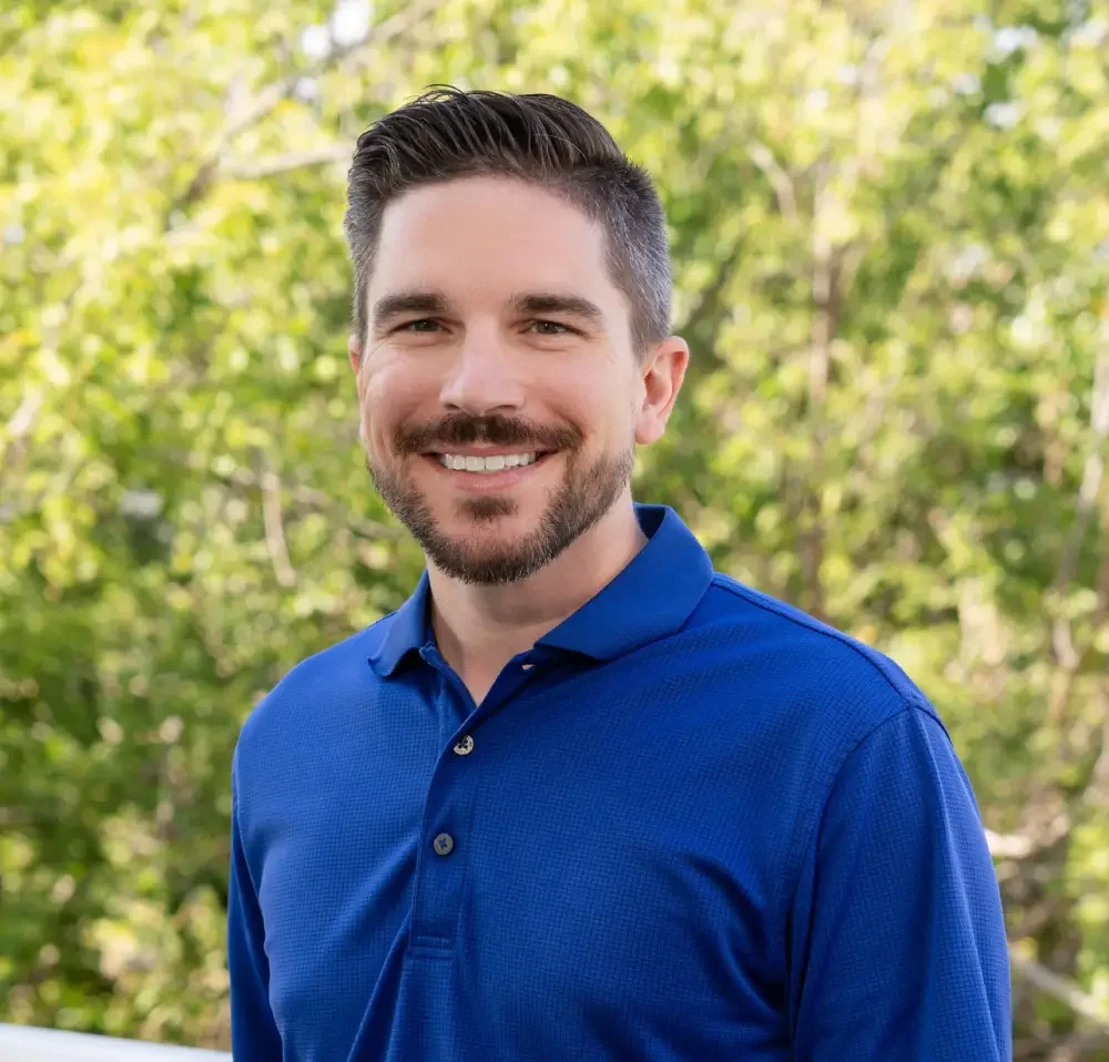 Smiling man with dark hair and beard wearing a blue polo shirt outdoors in a leafy setting