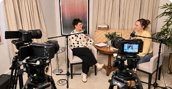 Two women recording a podcast or video interview in a cozy studio, filmed with multiple cameras and microphones.