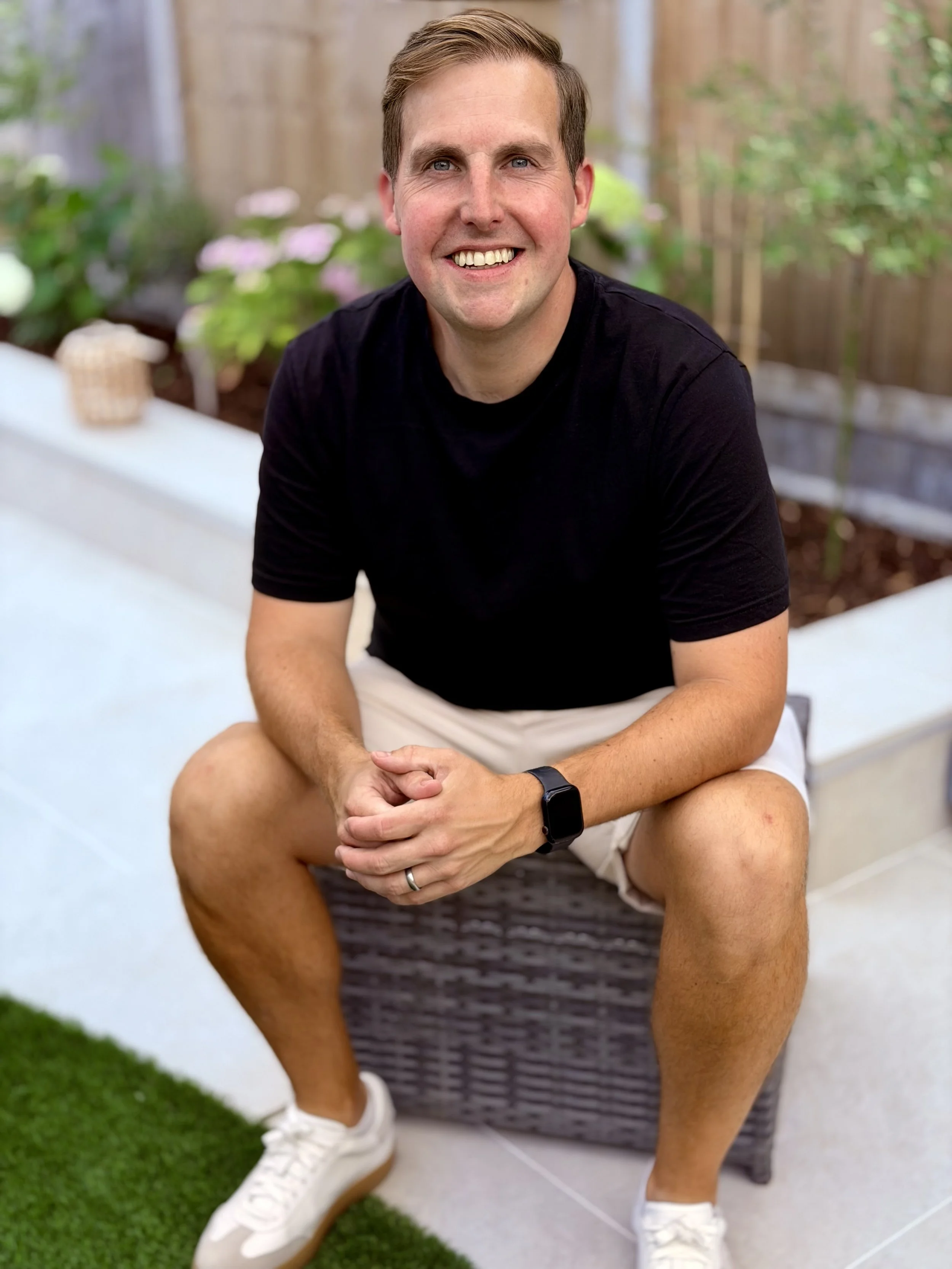 A man with short brown hair, blue eyes, and a broad smile, sitting outdoors on a wicker bench, wearing a black T-shirt, khaki shorts, white sneakers, and a smartwatch, with a garden and a wooden fence behind him.