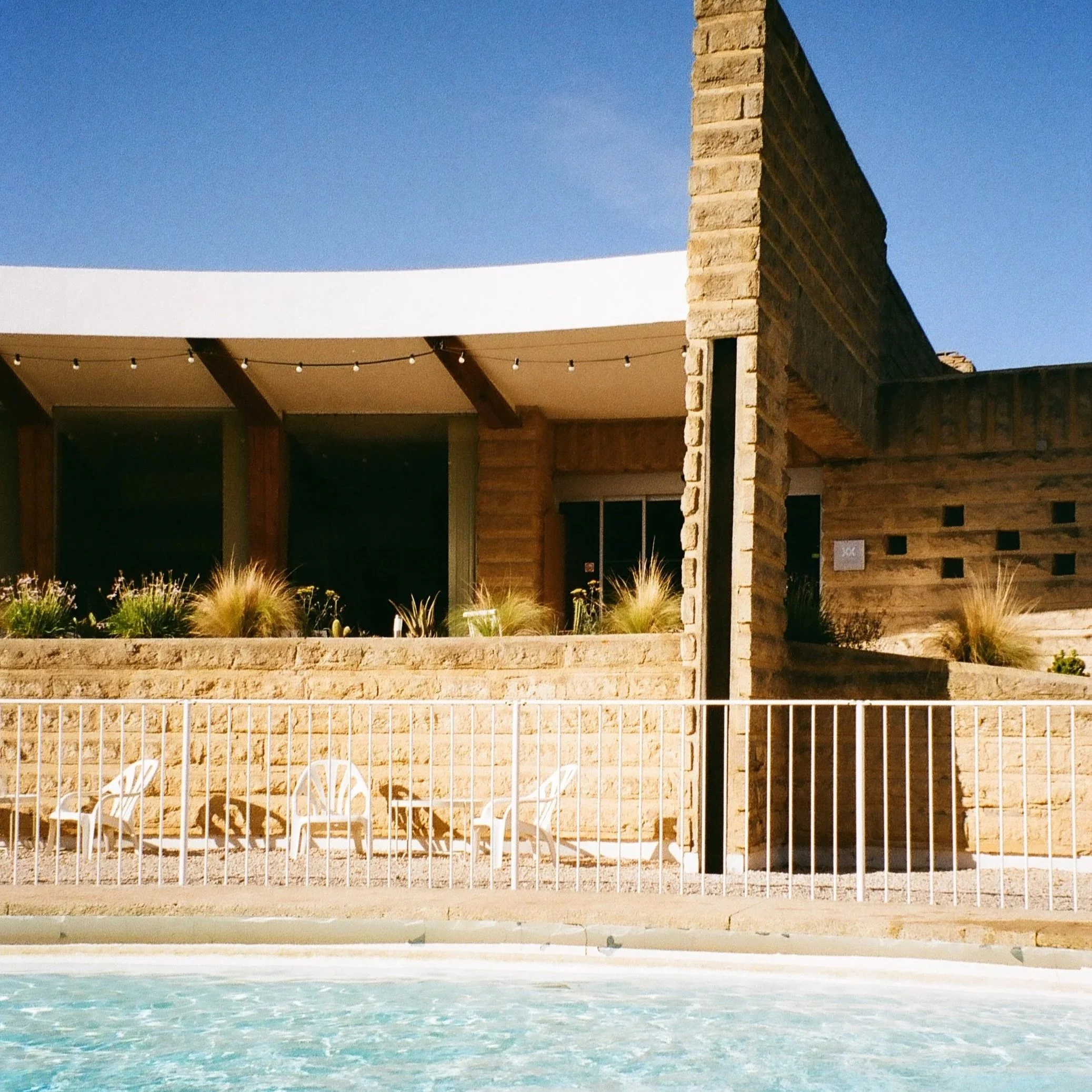 View of a backyard with a swimming pool in the foreground, a brick wall on the right side, a beige building with a balcony on the left, and a clear blue sky above at dusk.