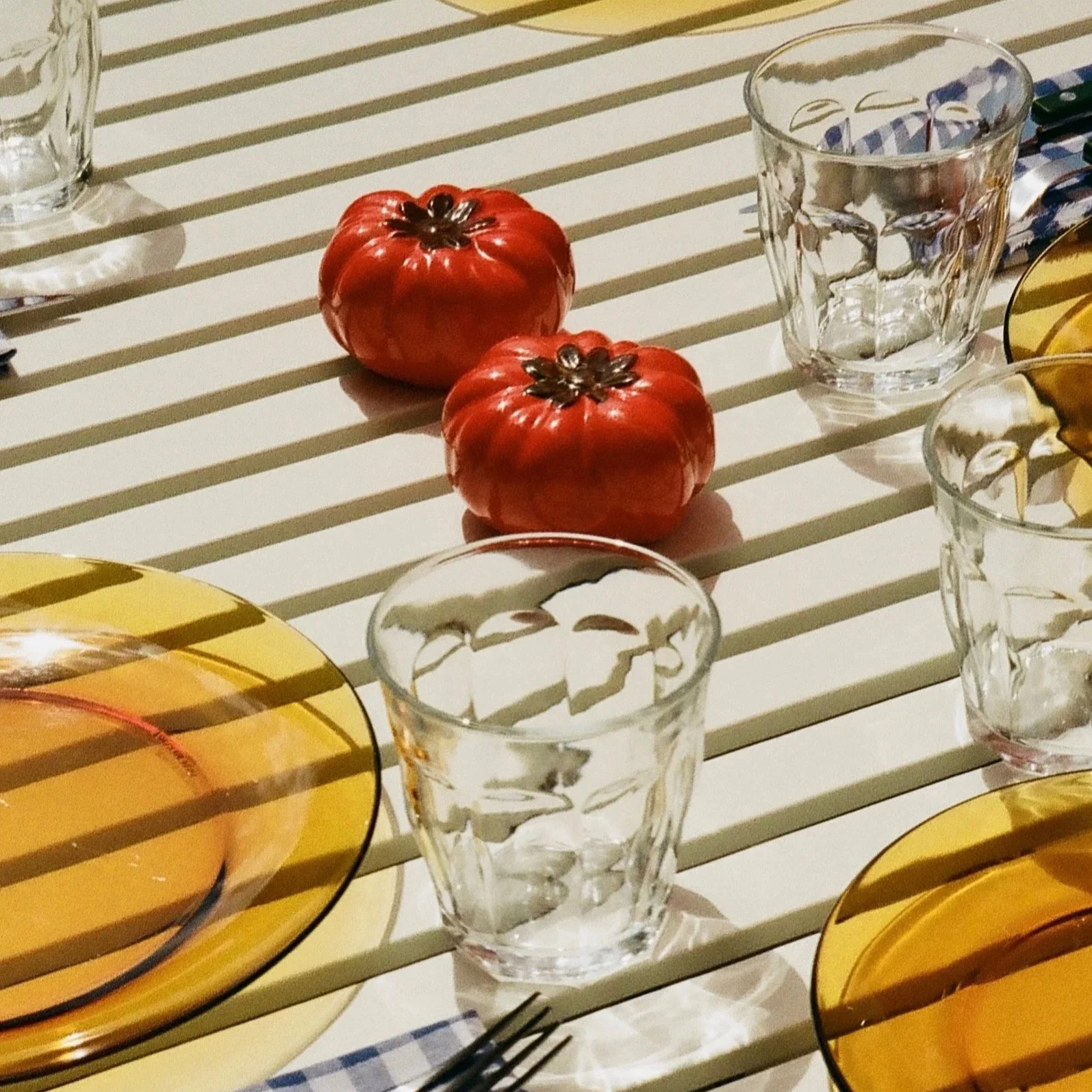 Reflection of a glass dining table with two red tomatoes, three empty crystal glasses, and yellow plates with striped tablecloth underneath.