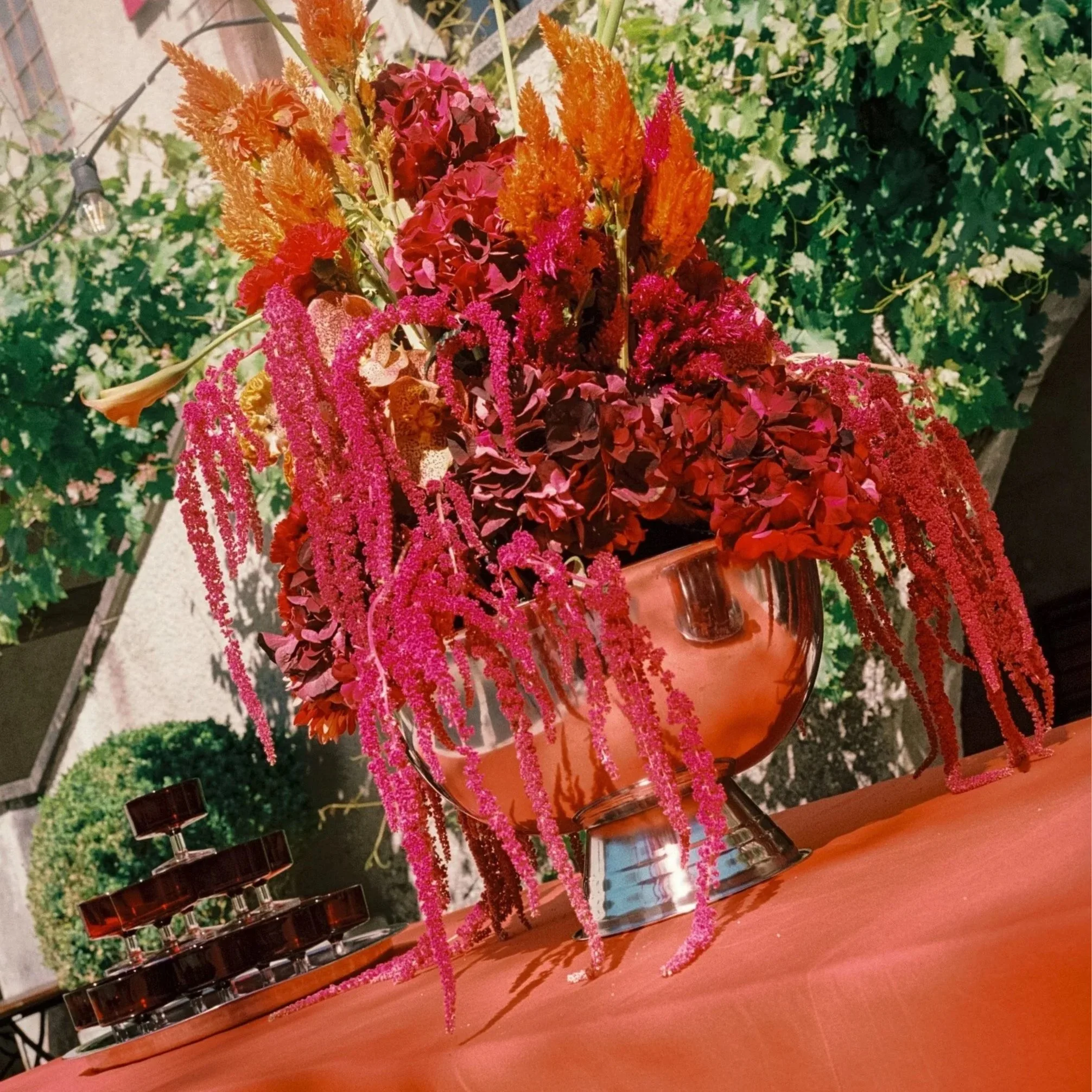 A floral display in a clear glass vase on a wooden surface with small bottles of red and black nail polish at the base. The arrangement includes red, pink, orange, and purple flowers and tall pink fuzzy and orange feathery plants, with a background of leafy green plants and decorative string lights.