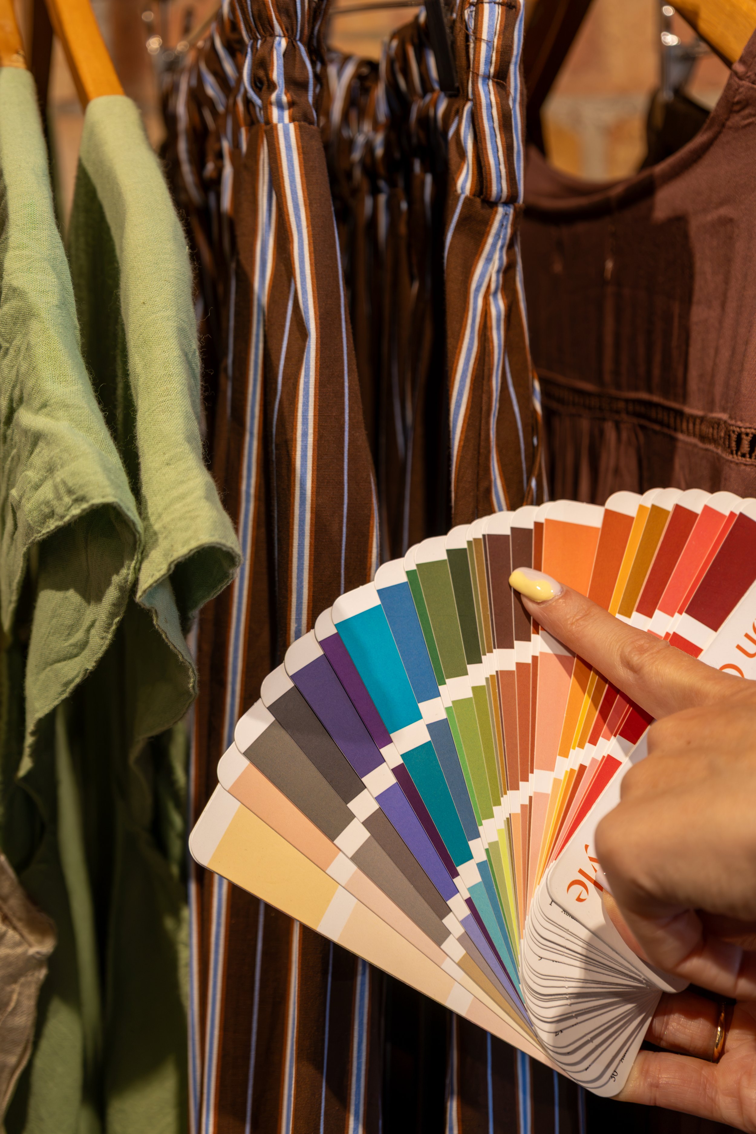 A person holding a color swatch fan deck in front of clothing hanging on a rack, including green, striped, and brown fabrics.