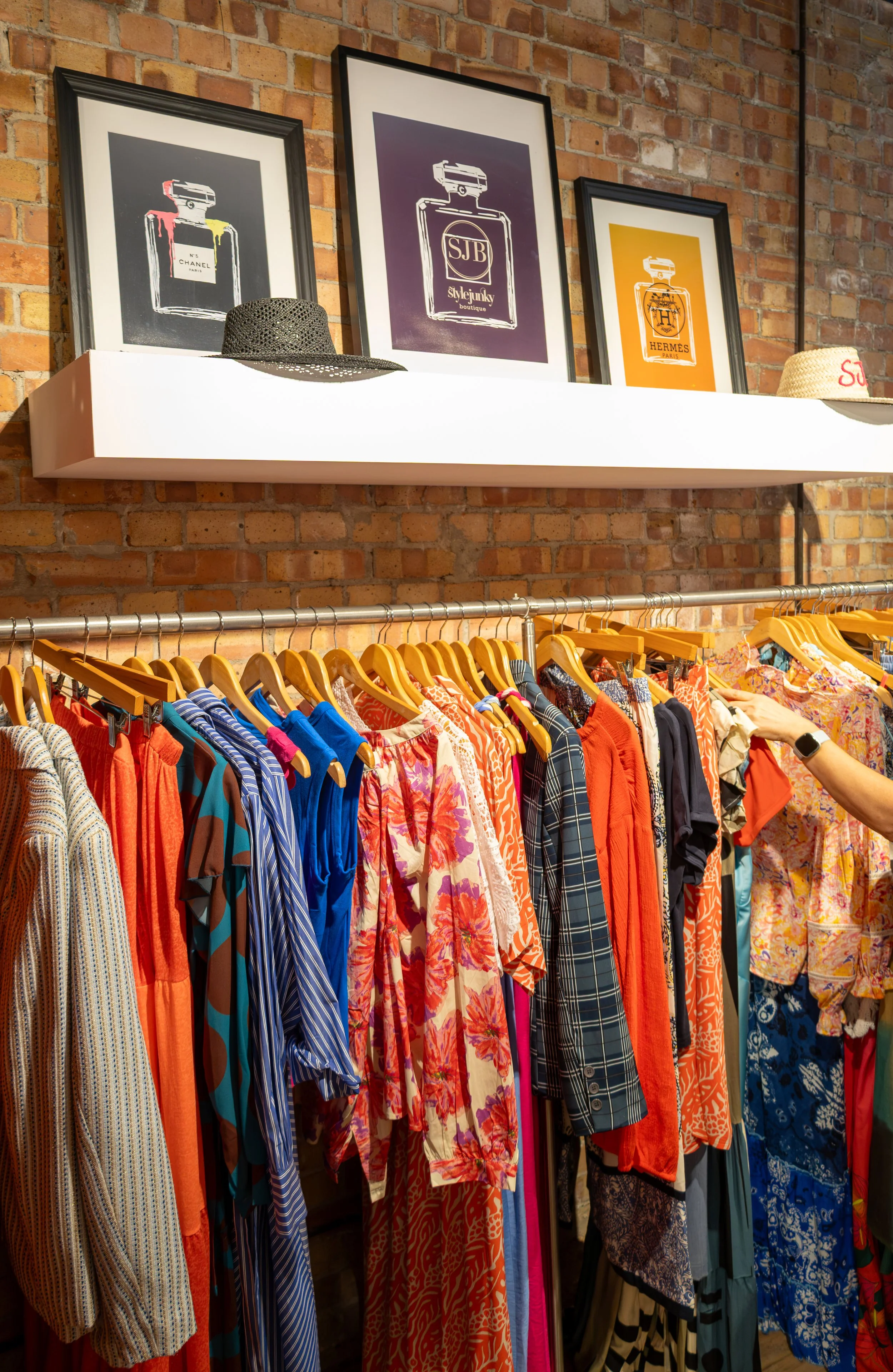 A clothing rack with colourful dresses and shirts, a white shelf with framed fashion artwork, and two hats placed on the shelf against a brick wall.