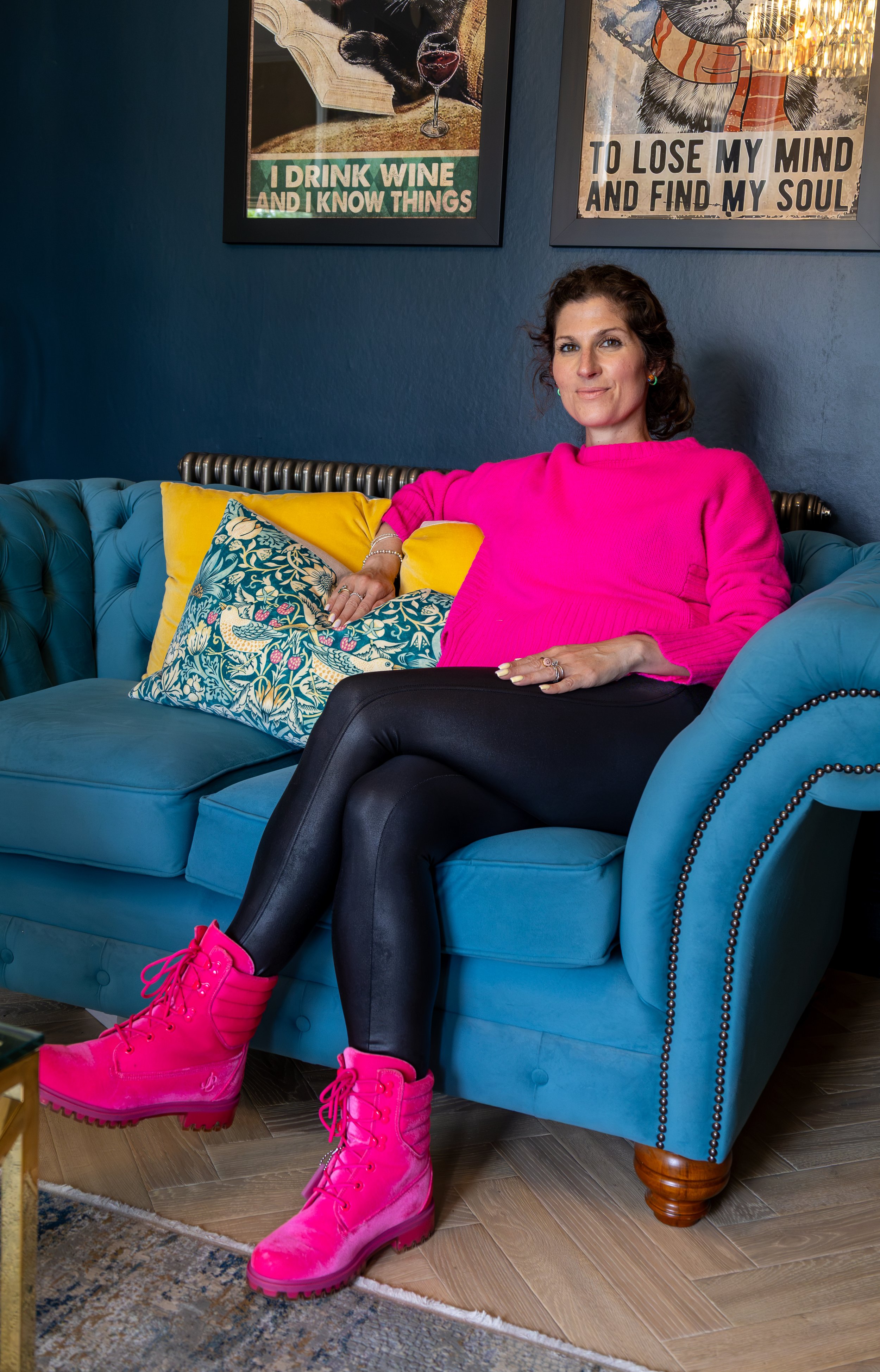 A woman sitting on a blue sofa with yellow and patterned pillows. She is wearing a bright pink sweatshirt, black leggings, and hot pink lace-up boots. She has earrings and a bracelet, with short curly brown hair and a slight smile.