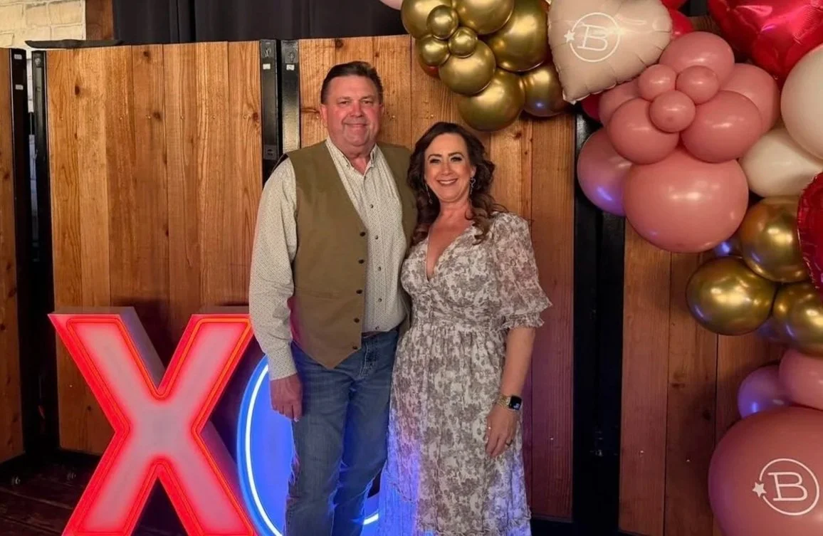 A man and woman stand together smiling in front of a wooden wall with colorful balloon decorations on the right side. The man wears a beige vest over a light shirt and jeans, while the woman wears a patterned dress. There is a large illuminated red letter "X" on the left side of the image.
