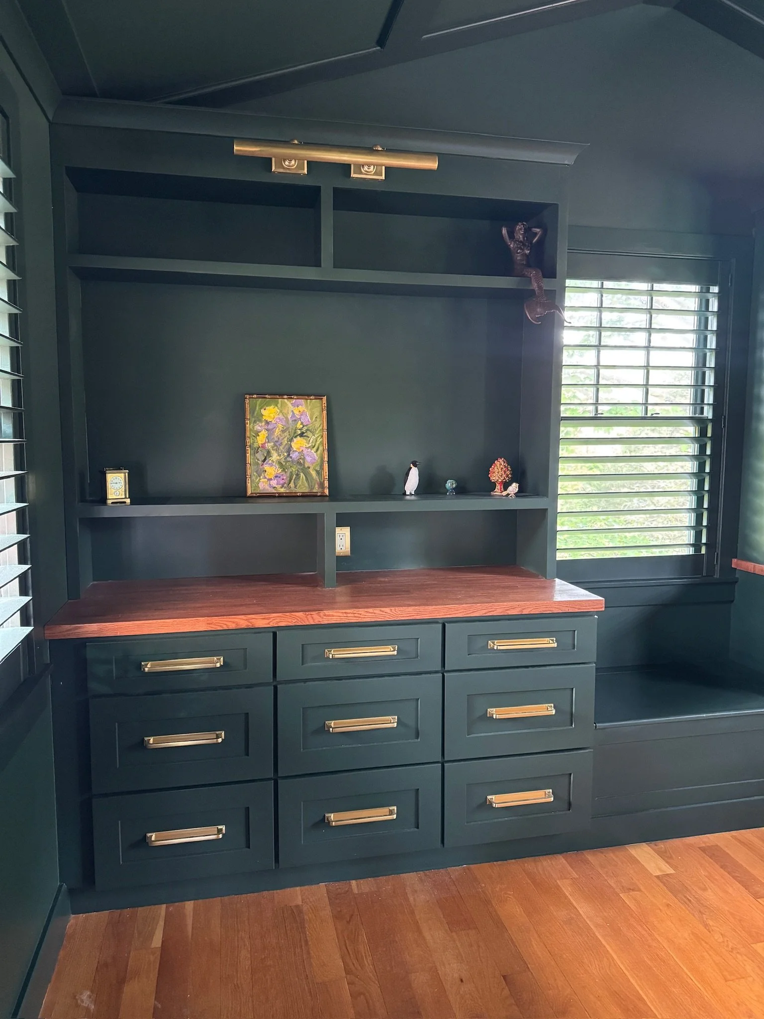 Dark green built-in bookshelf with gold handles, a wooden countertop, and small decorative items including a framed floral picture, small figurines, and a miniature tree, located by a window with horizontal blinds and hardwood flooring.