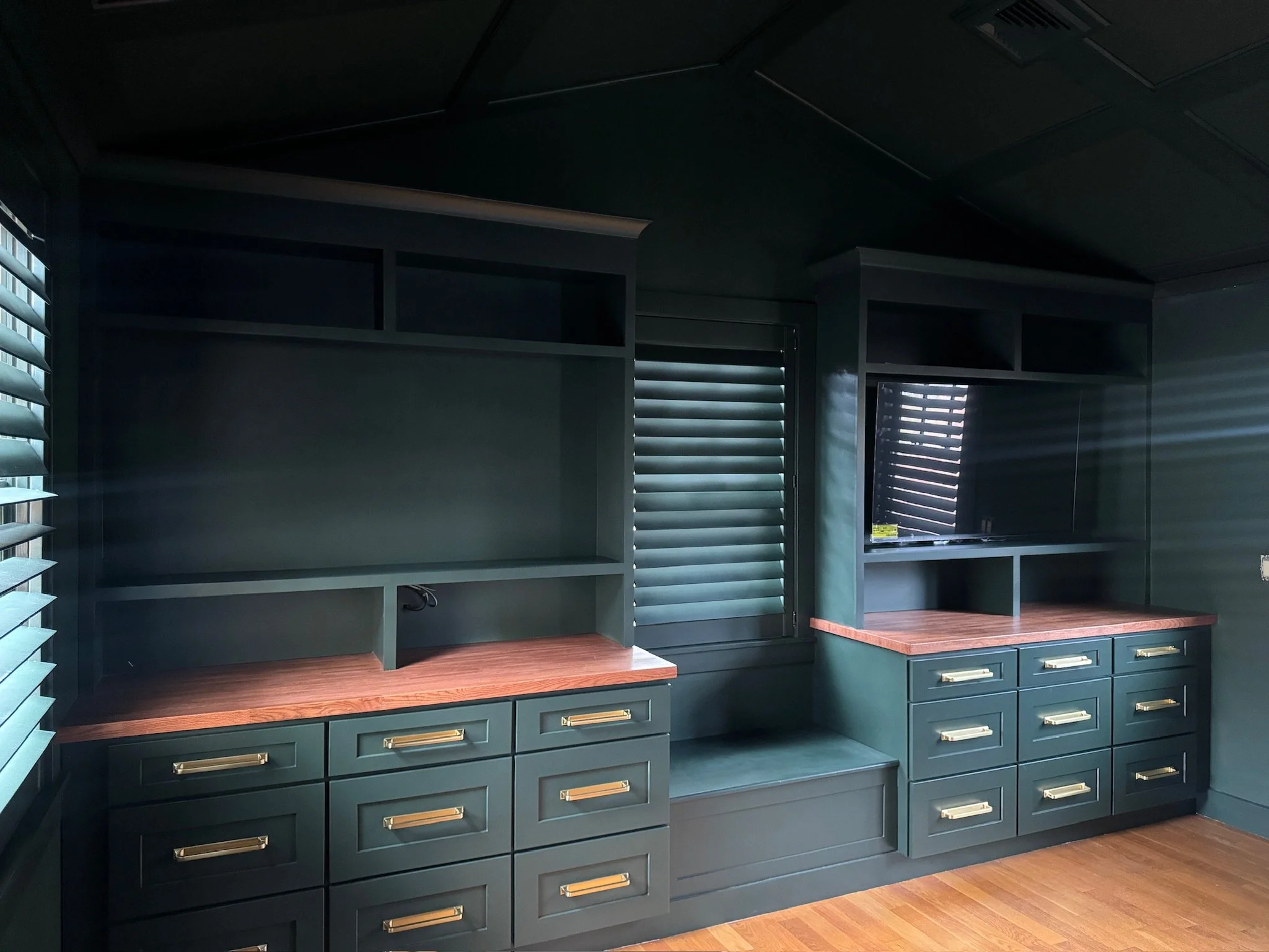 A dark teal kitchenette or similar room with multiple shelves, cabinets, and drawers, and two windows with horizontal blinds allowing some light to come through. The floors are wooden.