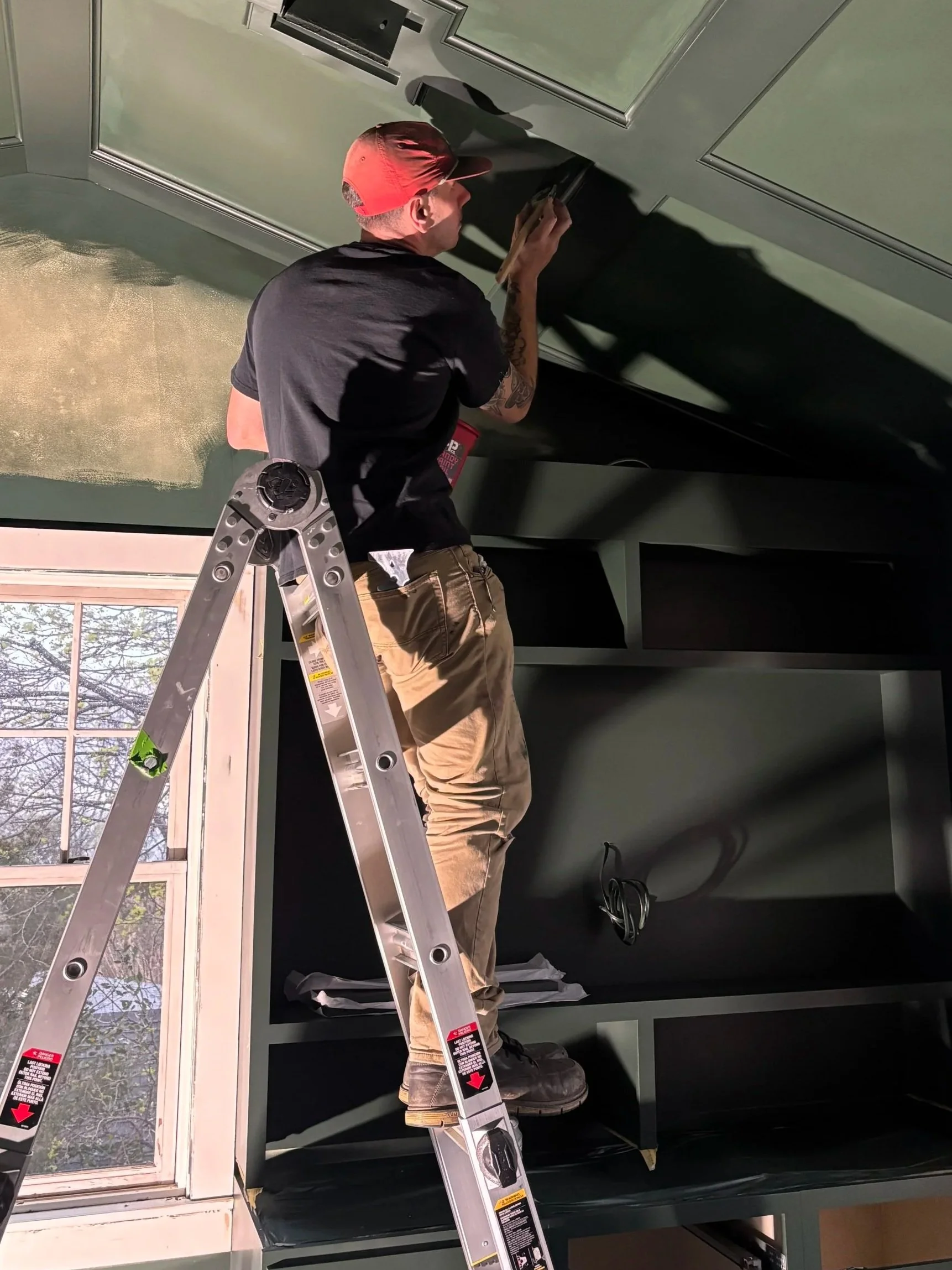 A man wearing a red cap, black t-shirt, tan pants, and black shoes stands on a ladder installing a cabinet or a wall panel inside a room. The room has a window with a view of trees and sky outside, and there are some wires visible near the floor.
