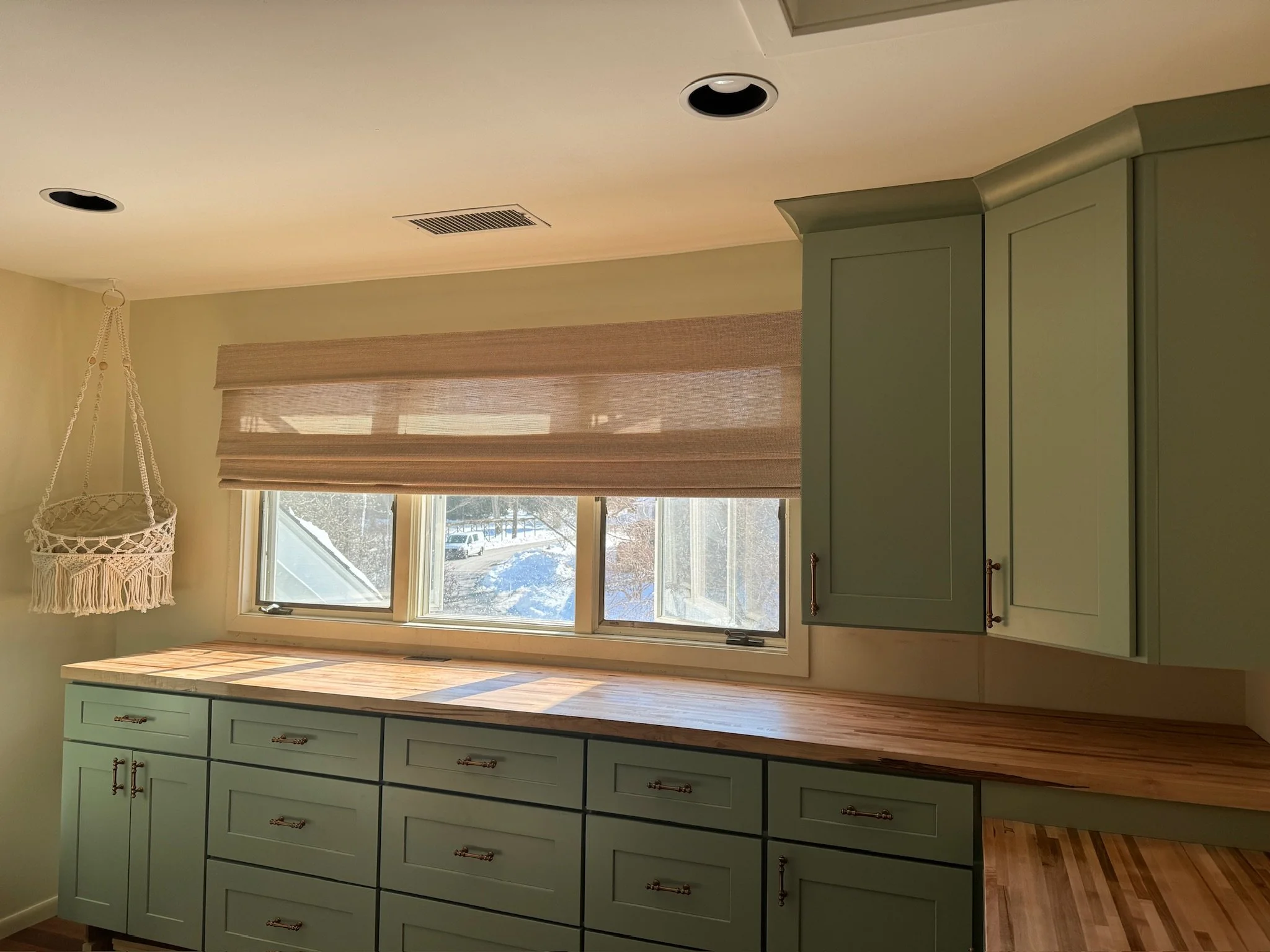A kitchen with green cabinets, a wood countertop, a window with beige blinds, and a hanging crocheted plant holder.