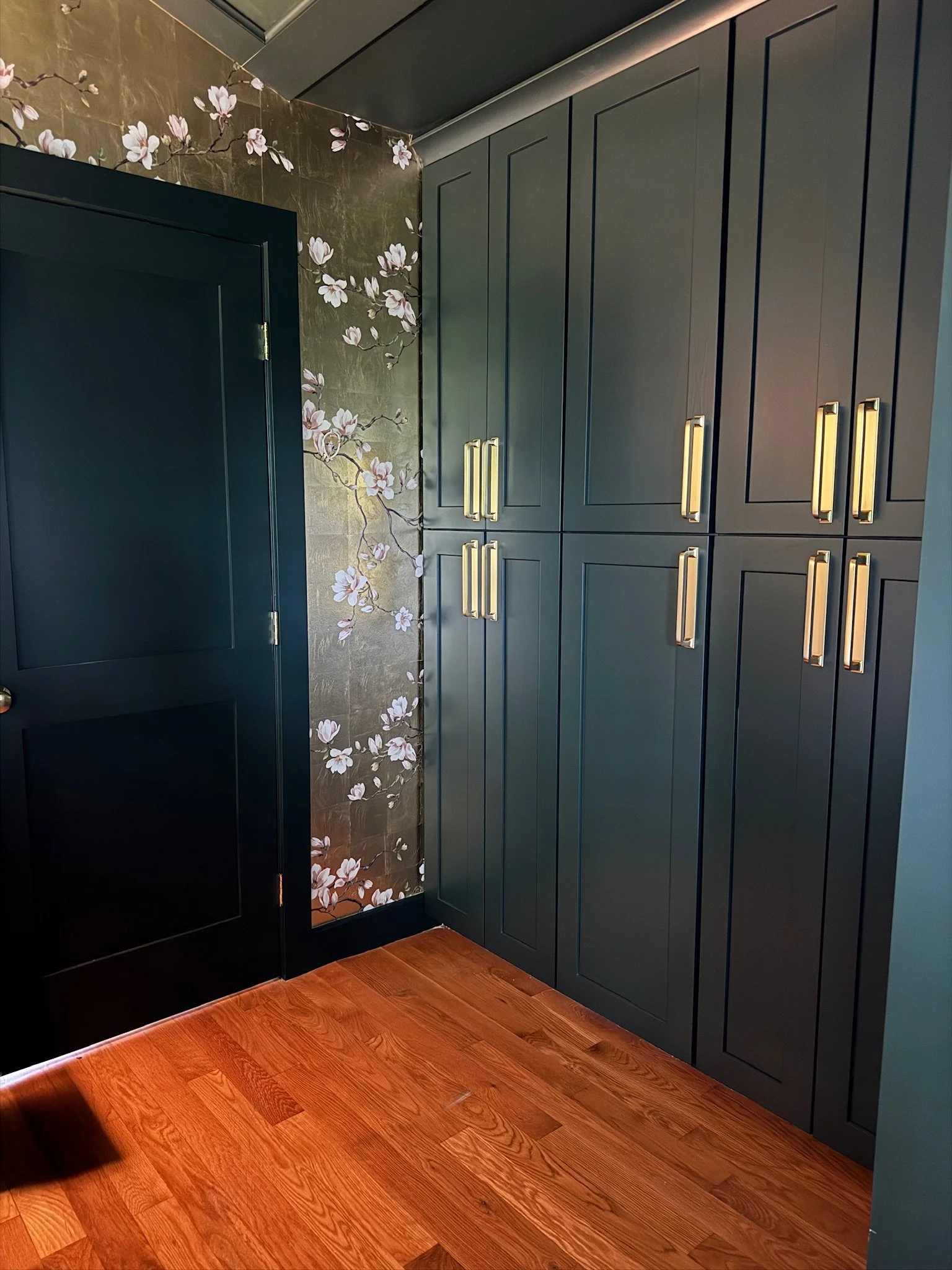 A corner of a room featuring dark painted cabinets with gold handles, floral wallpaper with pink and white flowers, oak flooring, and a closed door on the left side.