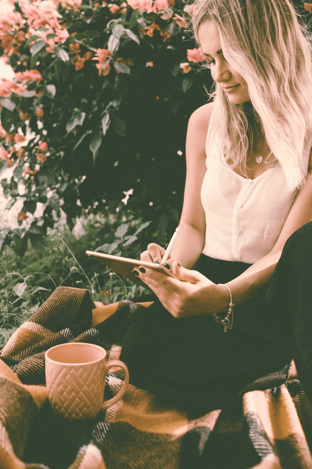 A young woman with blonde wavy hair sitting outdoors on a blanket, writing or drawing on a clipboard, with pink flowers and green foliage in the background. There is a beige mug nearby.