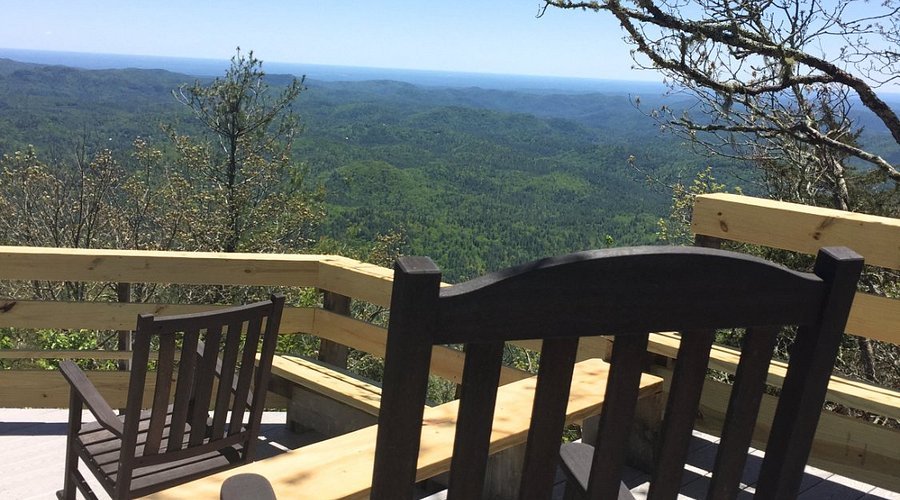 A scenic view from a wooden deck overlooking a forested landscape with rolling hills and distant mountains on a clear day.