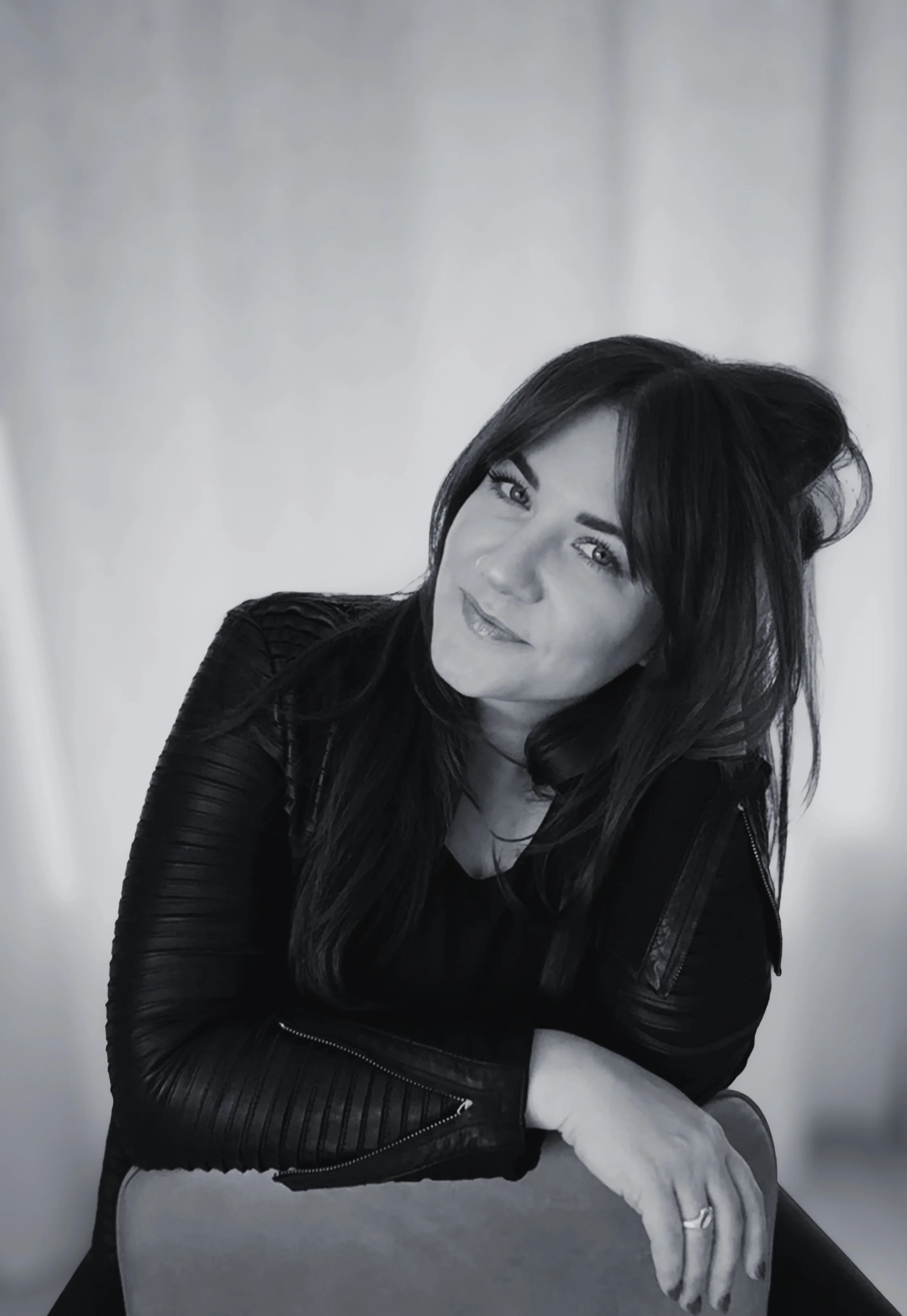 Black and white portrait of a woman with long dark hair, wearing a black leather jacket, seated with her head tilted and hand resting on a chair, gazing thoughtfully.