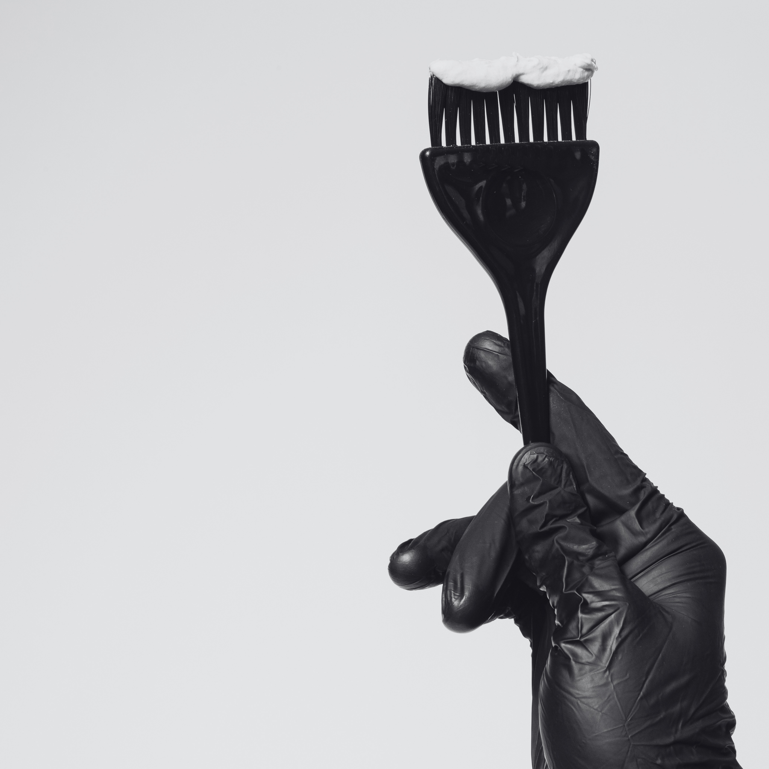 A person wearing a black glove is holding a black hairbrush with white lotion or cream on its bristles against a plain light grey background.