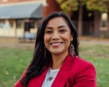 A woman with long black hair smiling outdoors, wearing a red blazer and earrings, with a house and trees in the background.