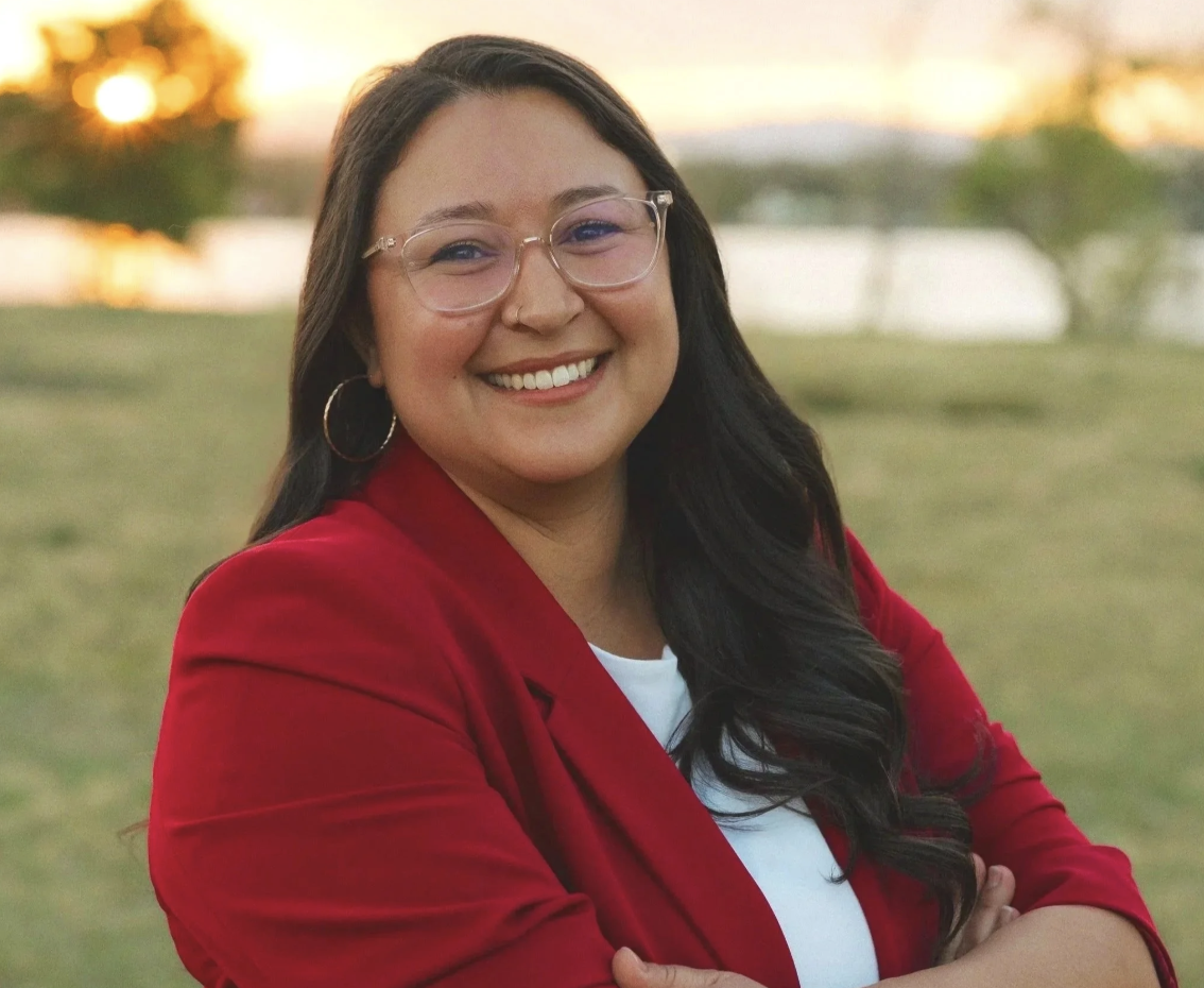 Smiling woman with glasses and hoop earrings in a red blazer outdoors at sunset.
