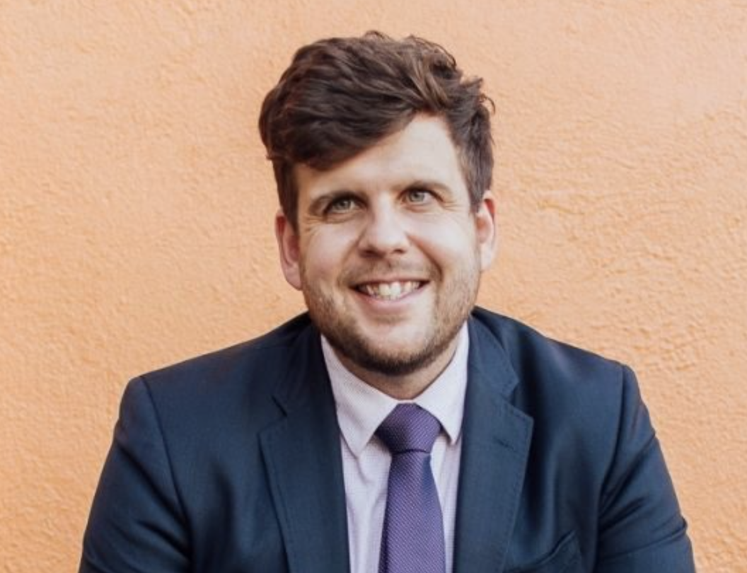 A smiling man with dark hair, wearing a navy suit, light shirt, and purple tie, sitting in front of an orange textured wall.
