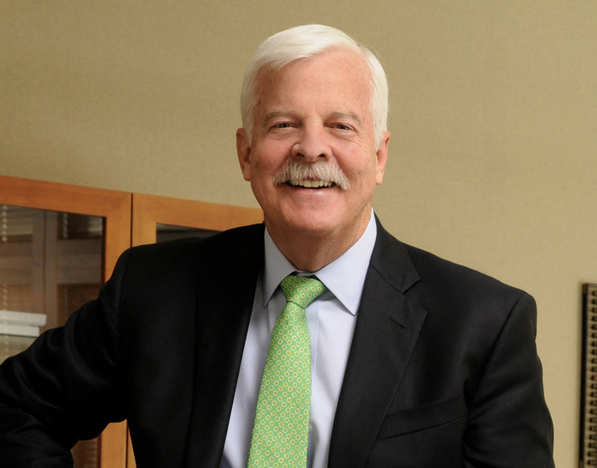 Senior man with white hair and mustache wearing a black suit, white shirt, and green patterned tie, smiling in an office setting.