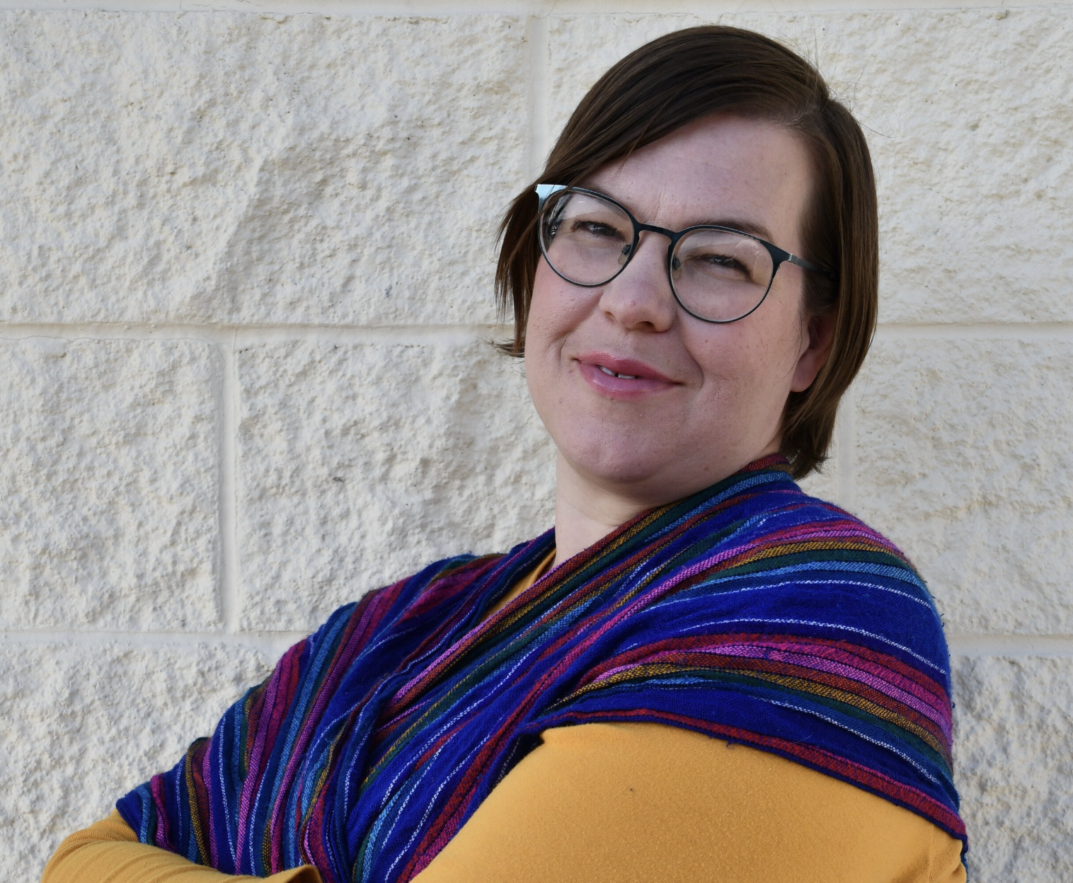 A woman with short brown hair, glasses, and a colorful scarf/wrap over yellow clothing smiling and standing against a textured beige wall.