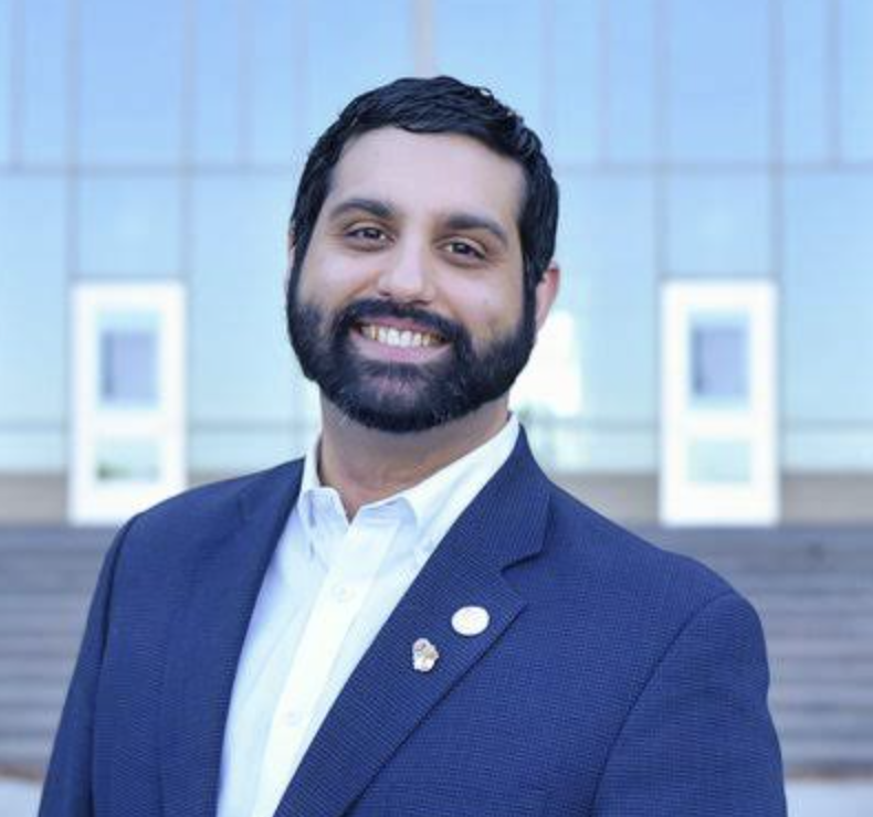 A smiling man with a beard and dark hair, wearing a navy blue suit and white shirt, standing outdoors in front of a modern glass building.