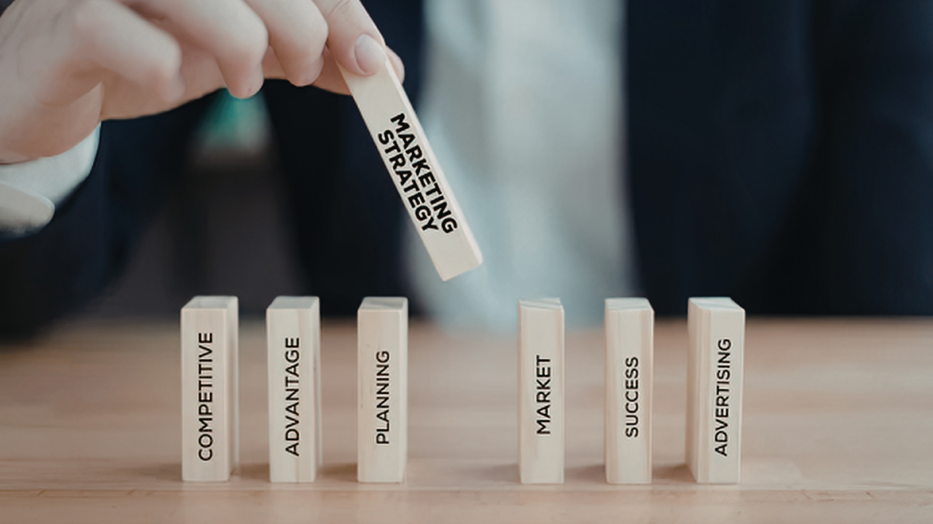 En hånd legger en liten blokk merket "Marketing Strategy" over andre blokker med ulike ord som "Competitive", "Advantage", "Planning", "Market", "Success" og "Advertising" på en bordflate.