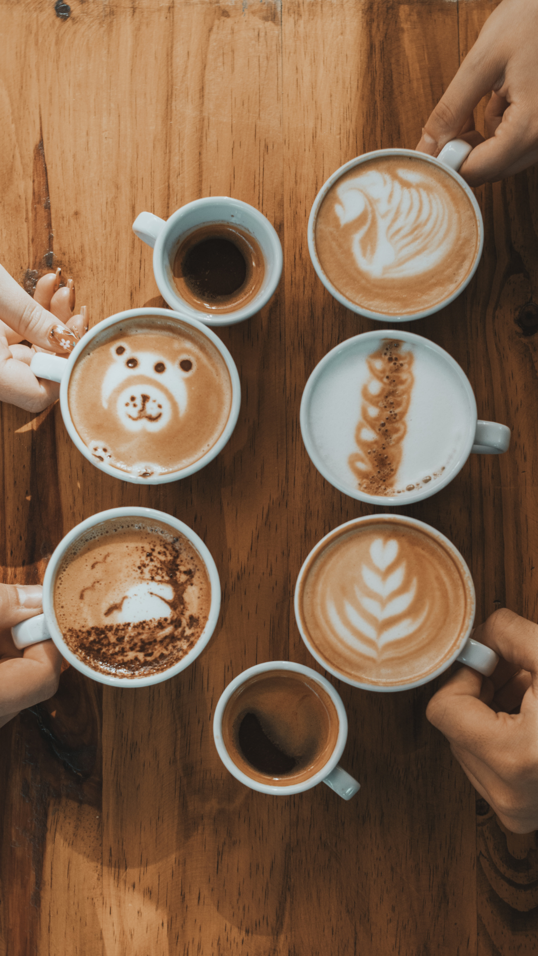 Fem kopper med kaffe, noen med lattekunst som skjelett, bear, hjerte og blad, på et trebord. Hendene til personer som holder koppene er synlige.