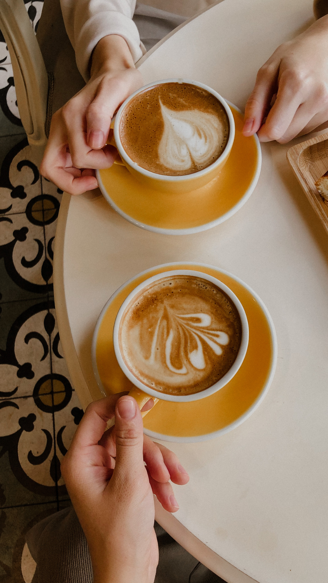 To kopper med cappuccino, hver med latte art, holdt av to personer over et rundt bord.