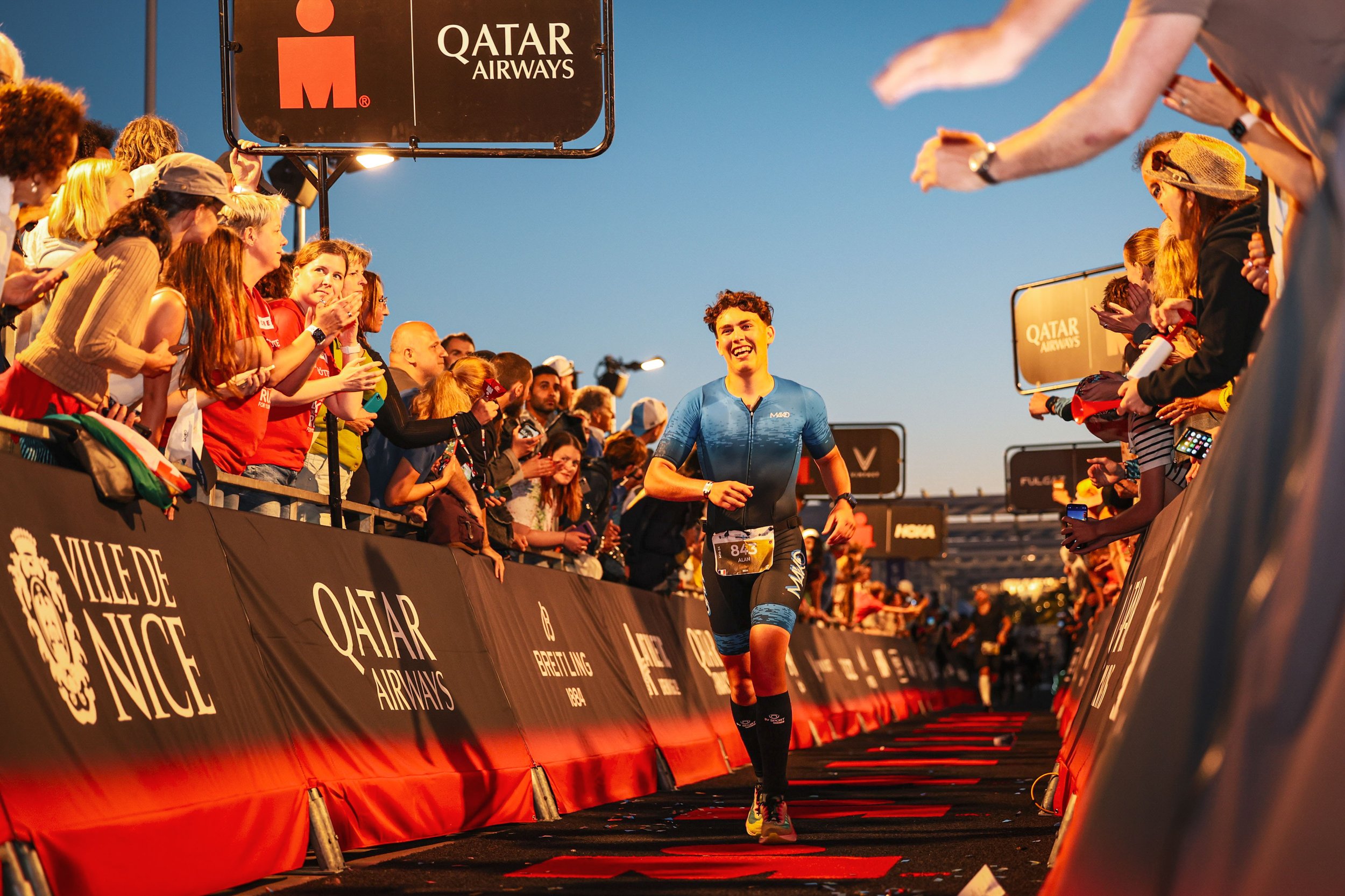 Un coureur traversant une ligne d'arrivée pendant un événement de marathon organisé par Qatar Airways, avec une foule d'admirateurs applaudissant et prenant des photos, et un ciel du crépuscule en arrière-plan.