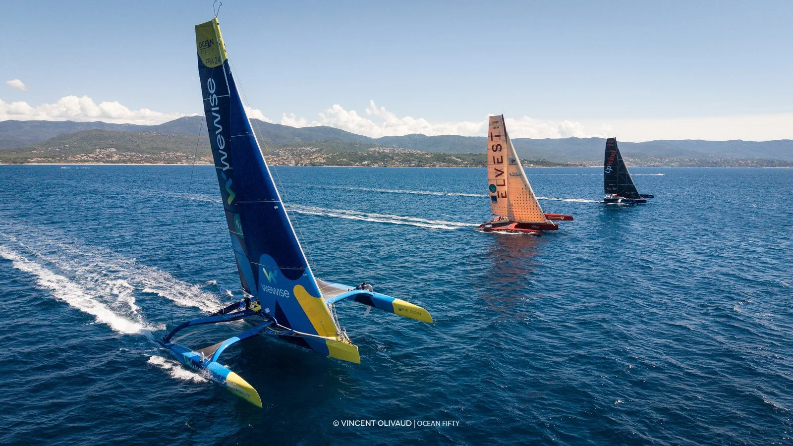 Trois hydrocars de course naviguant sur la mer, avec des montagnes en arrière-plan et un ciel ensoleillé.