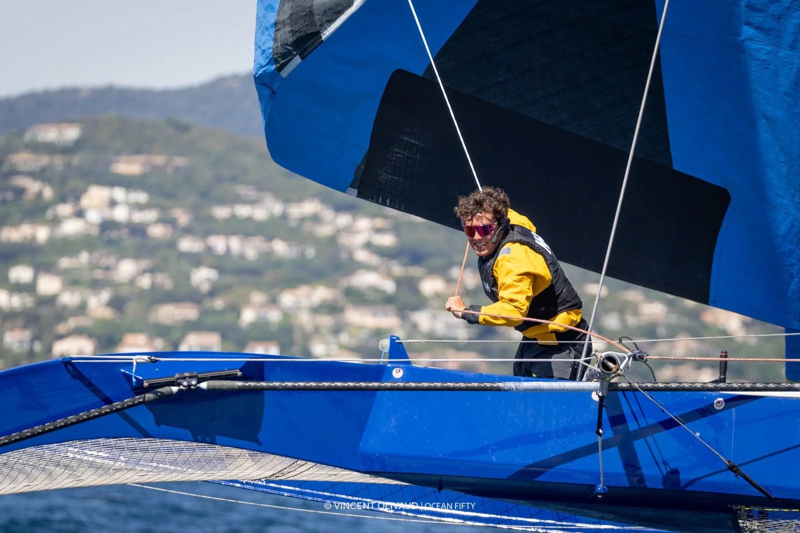 Voilier avec un marin portant des lunettes de soleil, un casque de communication, et une veste jaune, en pleine navigation près d'une côte montagneuse.
