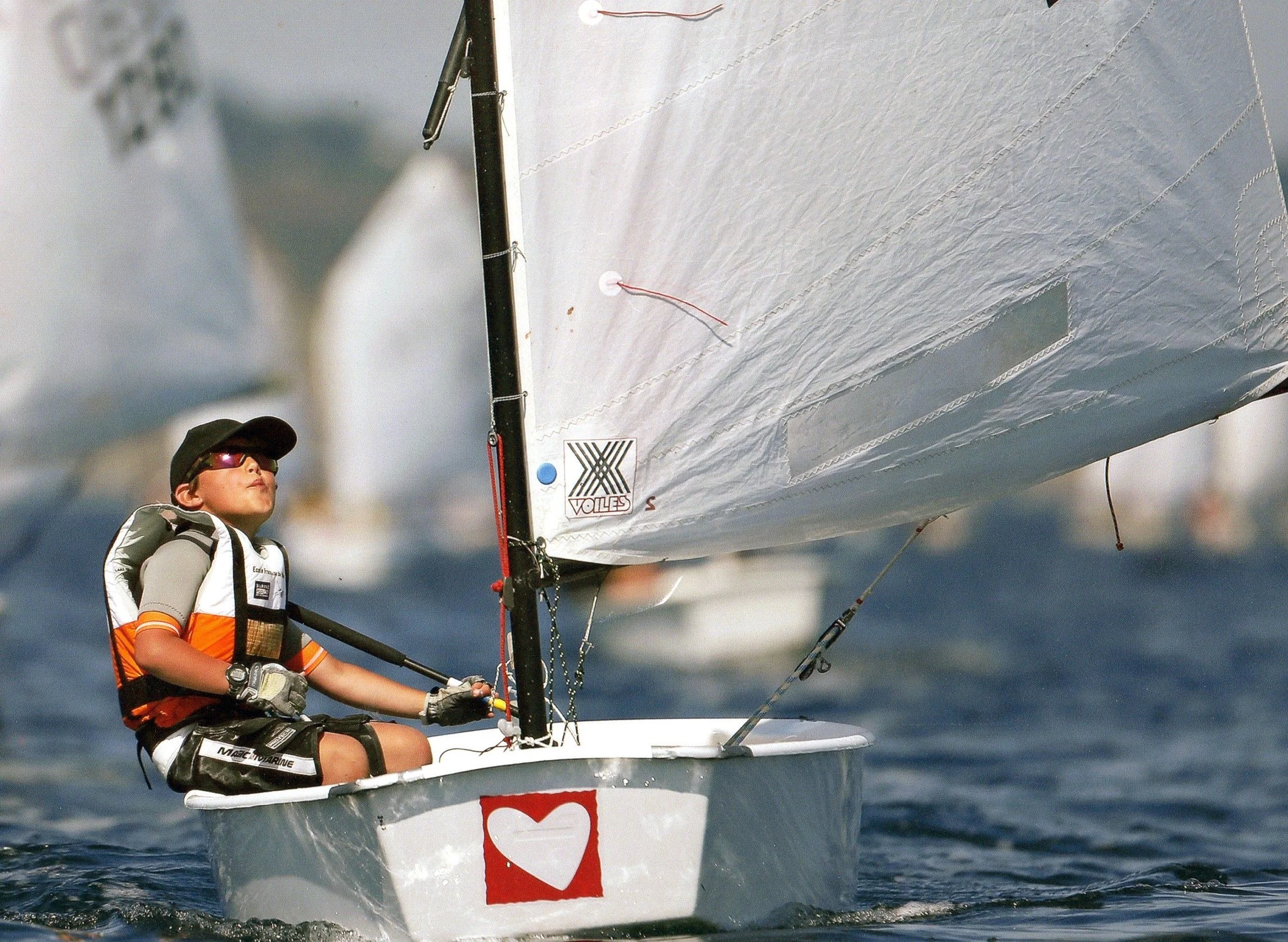 Jeune homme faisant de la voile sur l'eau, portant un équipement de sécurité et des lunettes de soleil, avec une voile de bateau à voile en toile blanche devant lui.