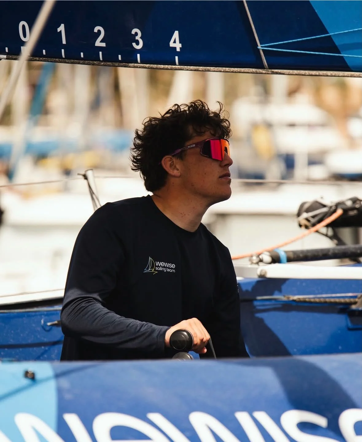 Jeune homme portant des lunettes de soleil réfléchissantes et un t-shirt noir avec le logo 'wewse sailing team' en bateau, à côté d'un bateau à voile dans un port.