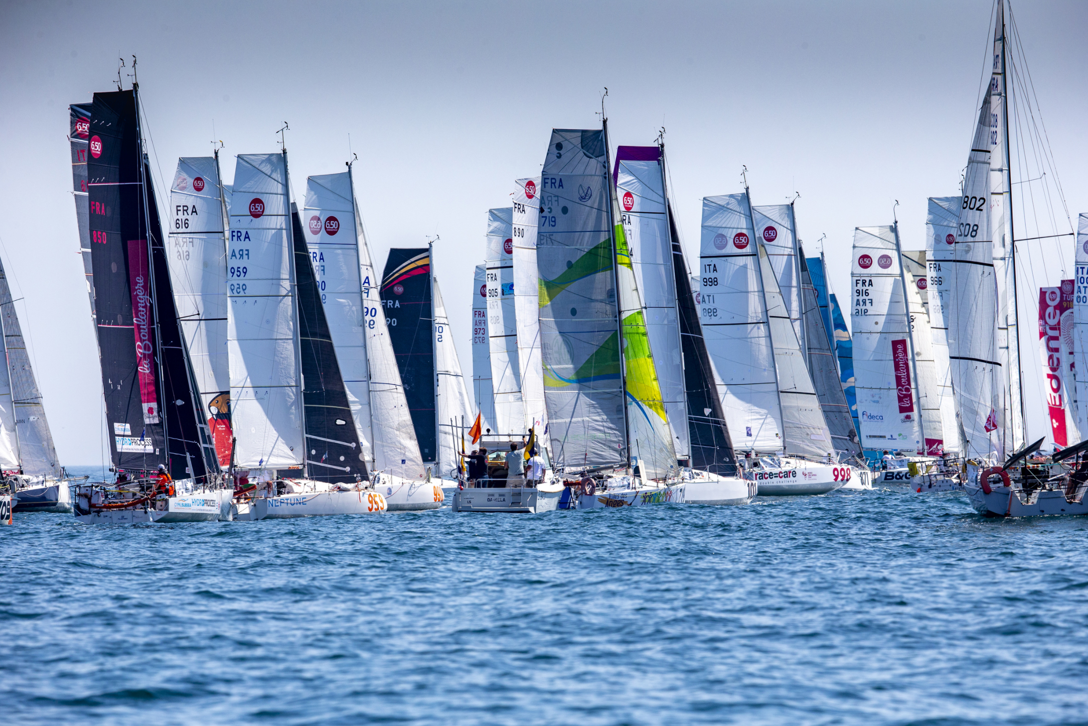 Fleets de voiliers en compétition sur l'eau, avec plusieurs voiles colorées et numérotées, lors d'une régate en mer.