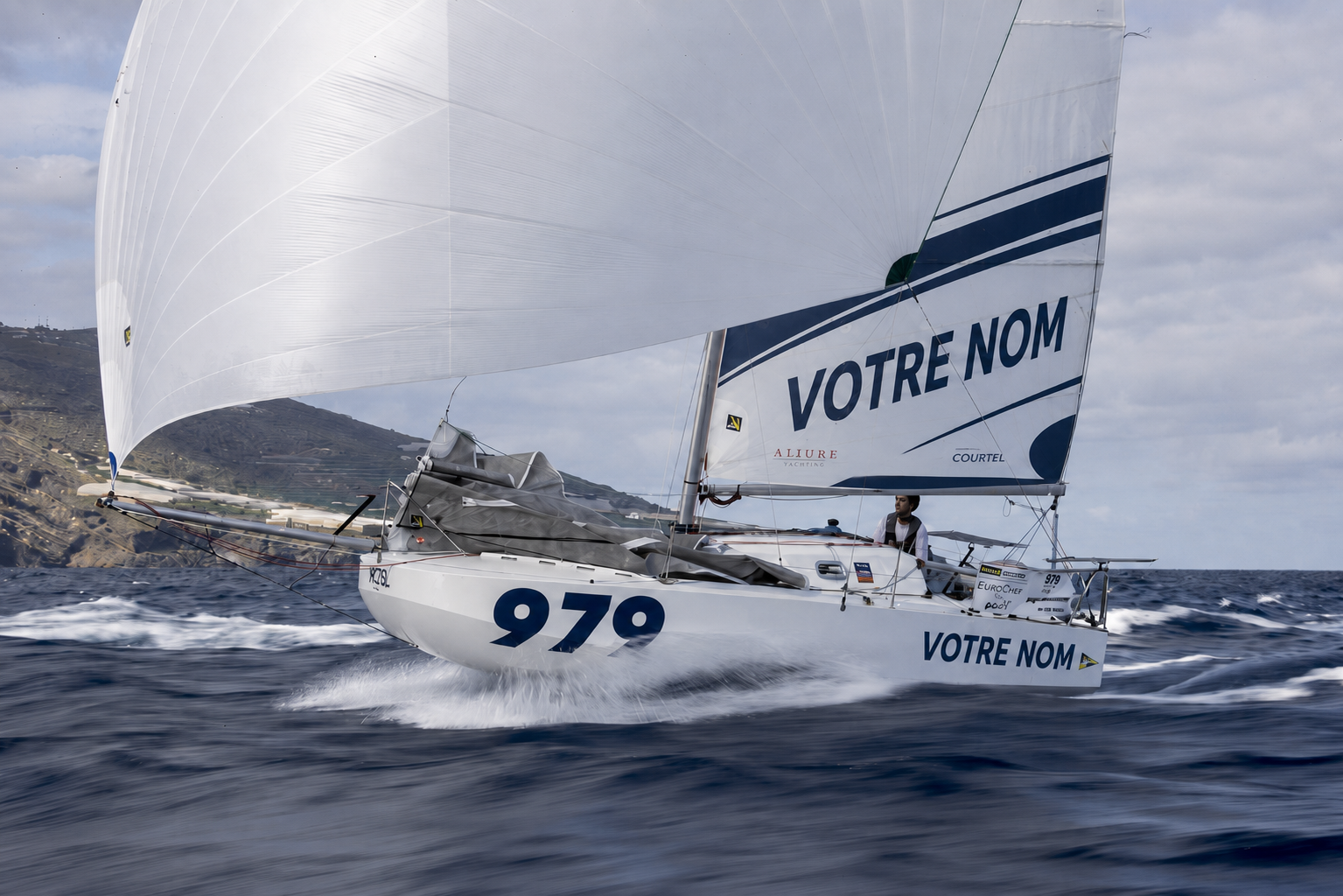 Un voilier blanc avec la numéro 979 naviguant en mer, portant le texte "VOTRE NOM" sur la voile et la coque, avec un arrière-plan de côtes et un ciel partiellement nuageux.